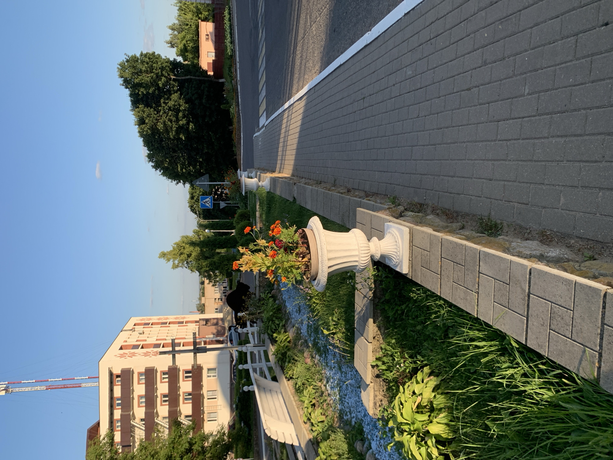 The image depicts a street scene in Kapyĺ, Belarus, bathed in the warm light of a late afternoon or early evening sun. A paved sidewalk, constructed from rectangular gray bricks, runs along the right side of the frame. A white stripe demarcates the edge of the sidewalk, separating it from the asphalt road. A pedestrian crossing is visible on the road, marked with white stripes.
To the left of the sidewalk, a low stone wall borders a planted area filled with lush green grass and various plants. Two ornate white planters with orange flowers are placed on the wall. A white wooden bench is situated further back in this area, beside a decorative dry streambed filled with small blue stones.
In the background, a multi-story building with a distinctive facade featuring repeated orange diamond patterns and brown balconies stands prominently. A tall radio tower with red and white sections rises into the clear blue sky behind the building. Trees, some deciduous and others evergreen, are scattered throughout the scene, adding to the urban greenery. A pedestrian crossing sign with a blue background and white figures is visible near the road. The overall impression is one of a well-maintained urban public space.