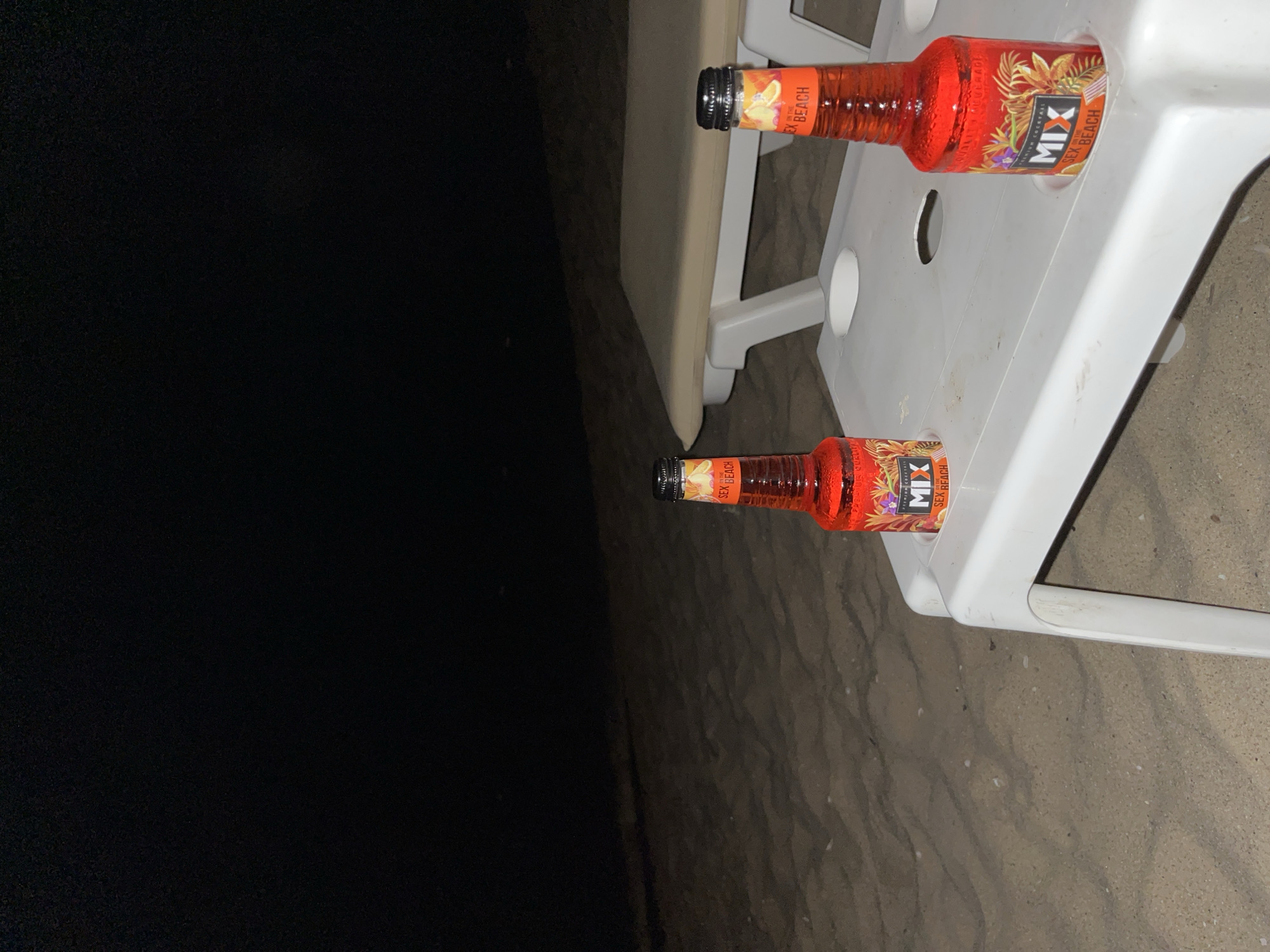 Two bottles of "Sex on the Beach" premium cocktail stand in cup holders on a white plastic table at night on a sandy beach in Obzor, Bulgaria. The bottles are red and have labels with images of palm leaves and flowers. The beach is dark, and a tan lounge chair is visible in the background. The sea is not visible.