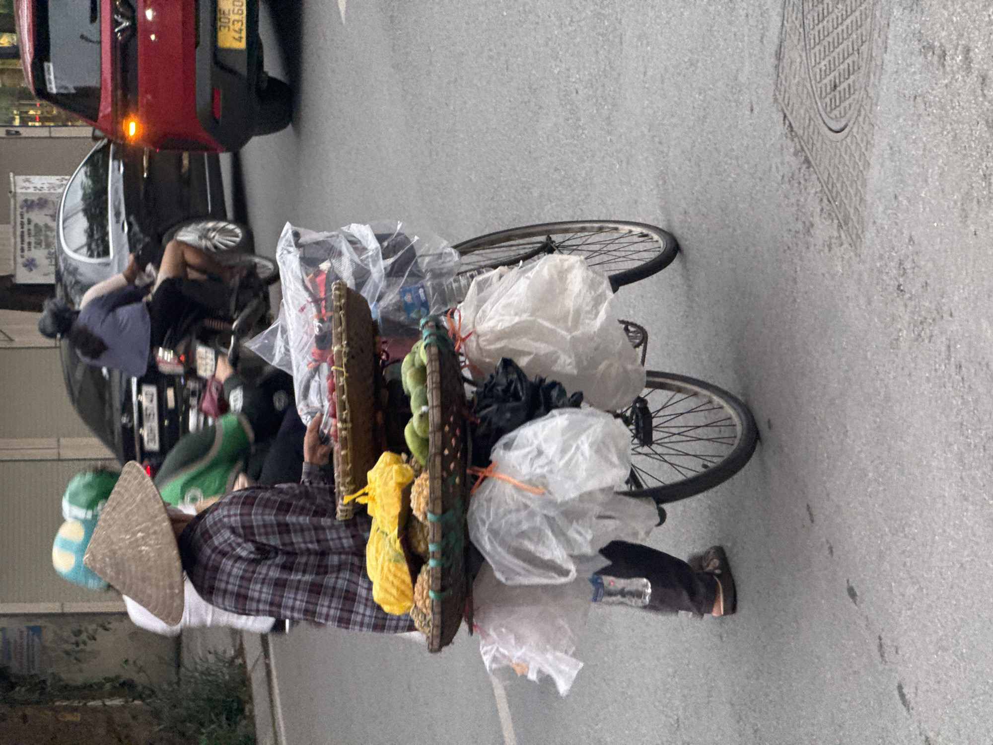 A street vendor with a conical hat pedals a bicycle through a street in Hà Nội, Vietnam. The bicycle is heavily laden with yellow and green produce, secured in baskets and plastic bags tied to the frame. The vendor wears a plaid shirt. Cars and motorcycles are parked along the side of the street. A manhole cover is visible on the road.