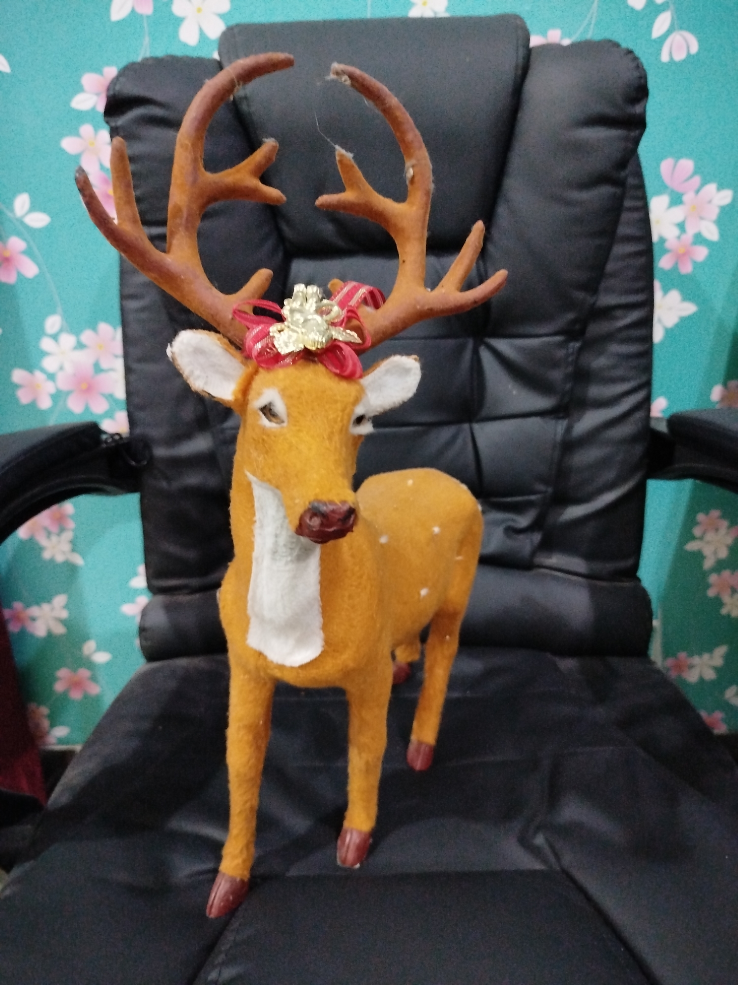 A stuffed reindeer Christmas decoration is placed on a black leather office chair. The reindeer is facing forward, with its head tilted slightly to the left. It has brown antlers, a white chest, and brown hooves. A red and gold bow is tied to its head. The chair is positioned in front of a turquoise wallpaper with a floral pattern of pink and white flowers. The scene is indoors, likely in a room within a residential or commercial building. There are no people present in the image. The lighting appears to be ambient indoor lighting, casting soft shadows. The image does not provide any specific clues about its location in Bandungrejo, Indonesia, beyond it being an indoor scene.