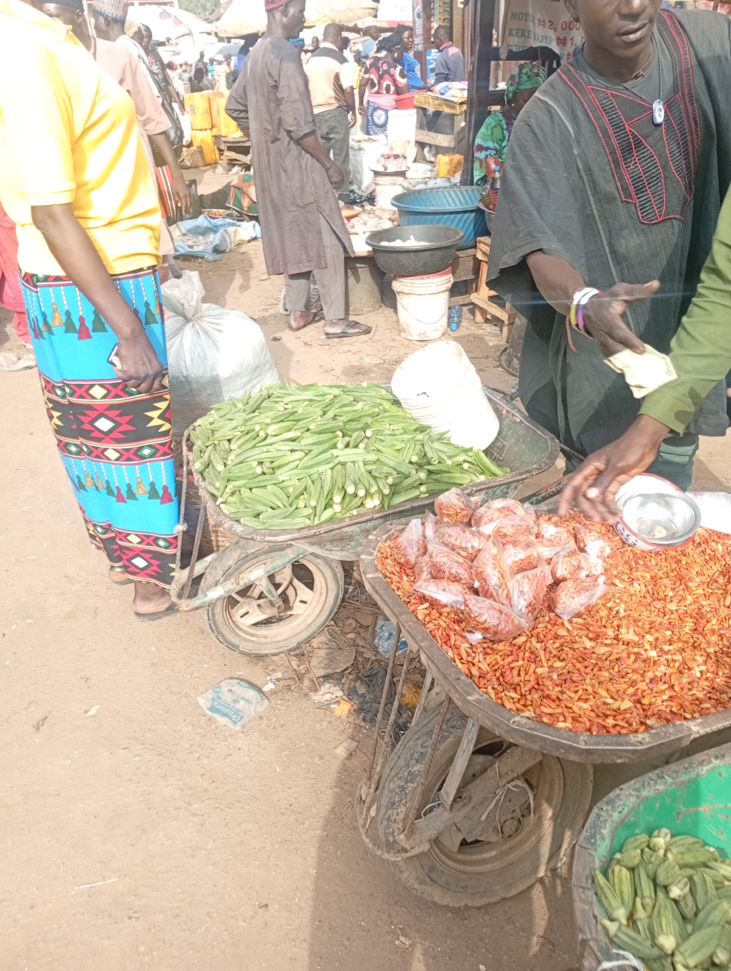 The image depicts a bustling outdoor market scene, likely in Rafin Par, Nigeria, characterized by vendors and stalls. In the foreground, a woman in a yellow shirt and a brightly patterned blue skirt stands next to a wheelbarrow filled with okra. To her right, a man in a dark, embroidered traditional garment appears to be making a transaction, holding what seems to be cash. Another wheelbarrow in the foreground is filled with dried red peppers and some smaller bags of what could be chili peppers or spices. Behind them, other market-goers and vendors are visible, along with various goods and containers, suggesting a lively commercial environment. The ground is dusty, indicative of an outdoor setting.