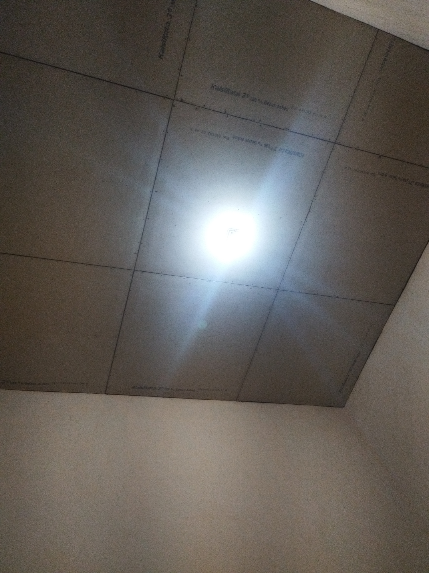 The image shows a ceiling constructed from large, light-colored panels that are joined together with visible seams. There are faint markings on the panels that appear to be text or product information. A bright light source is visible in the upper center of the image, casting a glare. The ceiling meets the walls of a room, which are painted a lighter shade of beige. The image provides no information about the city of Kalidawir, Indonesia, or any other location. There are no people or actions depicted in the image.