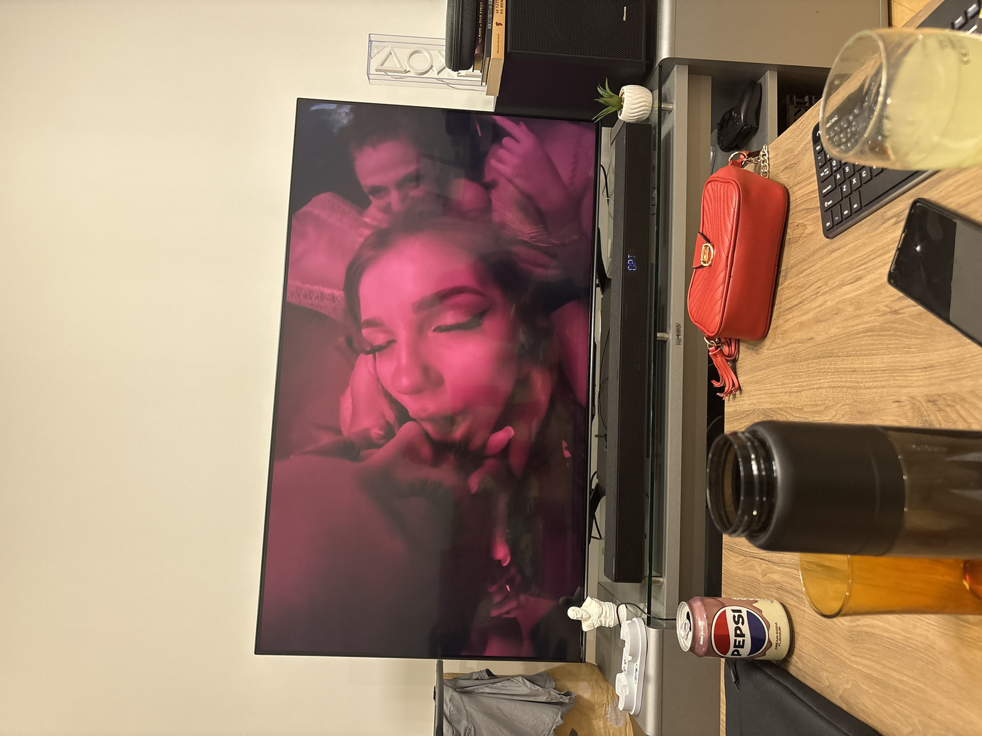 The image shows a television set with an amorous couple displayed on its screen. The television is placed on a light wood-grain desk surface. Other items on the desk include a computer keyboard, mouse, a black smartphone, a red purse, a half-full glass of liquid, a bottle with a black cap, and a can of Pepsi Cream Soda Zero. A sound bar is positioned in front of the television. A small white planter with a green plant sits on the shelf above the soundbar. A set of books and a black speaker are visible above the television. A white PlayStation decoration is affixed to the wall. Below the television, there is a charging dock and a gray t-shirt. The wall behind the television is a neutral beige color. The location is likely indoors and the exact location of the city of Lovech, Bulgaria, cannot be determined from the image.