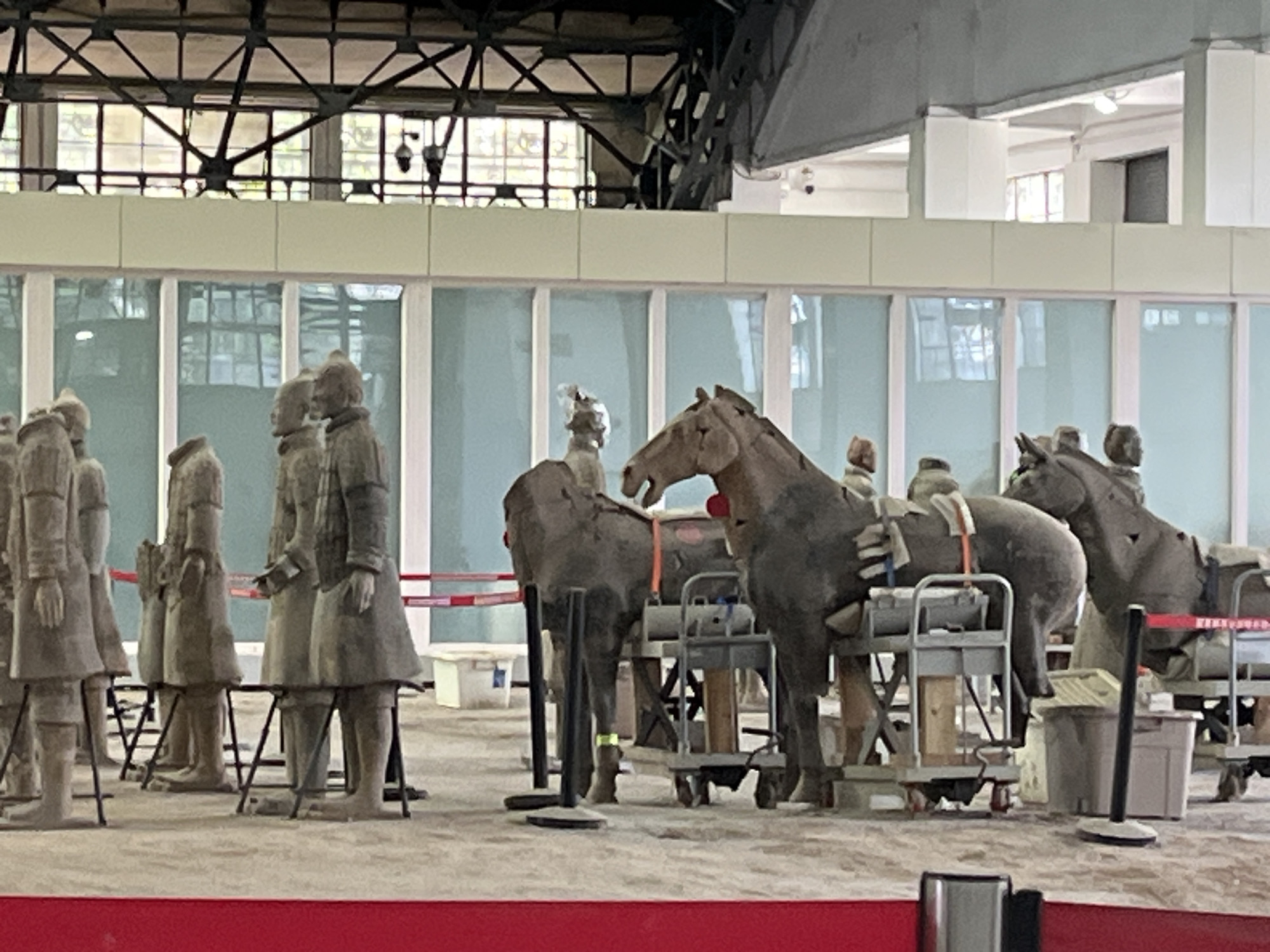 The image depicts a display of Terracotta Army figures and horses within a museum-like structure. In the foreground, several life-sized terracotta soldier figures are standing, partially obscured by black metal stands. Further back, a procession of terracotta horses, some equipped with saddles and harnesses, are positioned on wheeled carts and display stands. The background reveals a modern building with large windows, structural beams, and industrial lighting, suggesting an indoor exhibition space. The overall scene is static, showcasing historical artifacts within a contemporary setting. The location is identified as Unknown, Unknown.