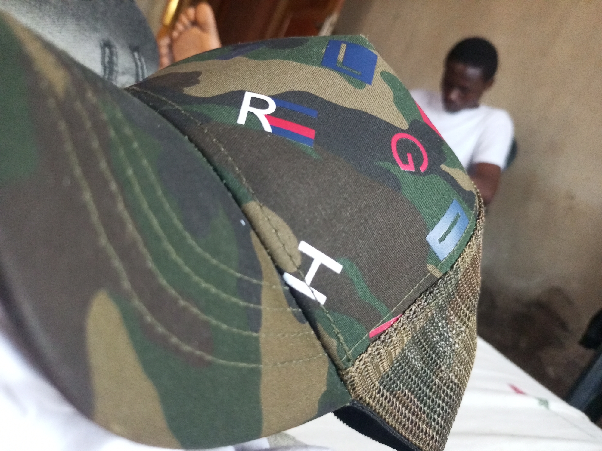 The image shows a close-up of a camouflage baseball cap with various letters and logos on it. The cap is in the foreground, with a blurred background that includes a person sitting down and a hint of feet. The cap features a letter "R" with red and blue stripes, a stylized pink "G", and a blue rectangle with a white border. There is also a white letter "H". The background suggests an indoor setting, possibly a room with plain walls. The lighting appears to be natural, indicating daytime. The person in the background is wearing a white t-shirt and their head is bowed, giving a sense of casualness or perhaps contemplation. The focus on the cap suggests it might be the primary object of interest, possibly indicating a personal item or an advertisement. The overall mood is informal and candid.