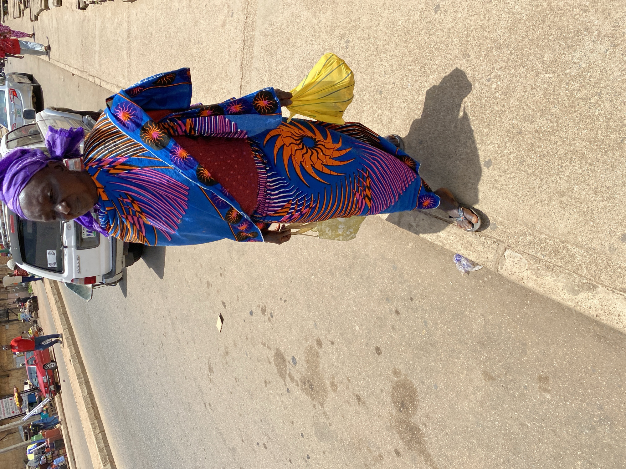 An elderly African woman stands near a busy Bukuru, Nigeria, street. She is wearing a traditional, brightly patterned blue dress with orange and purple details and a purple headscarf. She holds a yellow object, possibly a bag or cloth. The street shows signs of vehicle traffic and roadside commerce. A white van is parked nearby. The overall lighting suggests a sunny day.