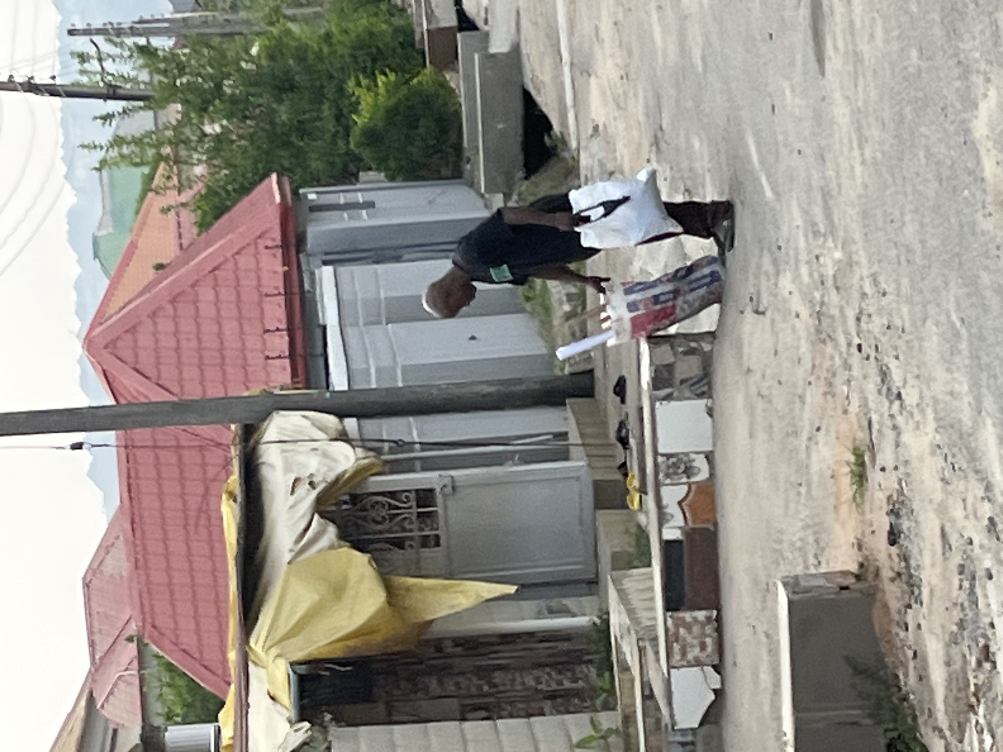 A man wearing a black t-shirt and light-colored shorts is bending over while standing on a street or sidewalk. He appears to be holding or placing something on a low wall or structure constructed from tiles. A white bag is near him, and an object with an image of a flag is on the ground close by. Buildings with red-tiled roofs and light-colored facades are in the background, along with a utility pole. The location is residential and appears to be a lower-income area. While there is no definitive sign that this is in Abuja, Nigeria, the housing style is consistent with residential areas there.