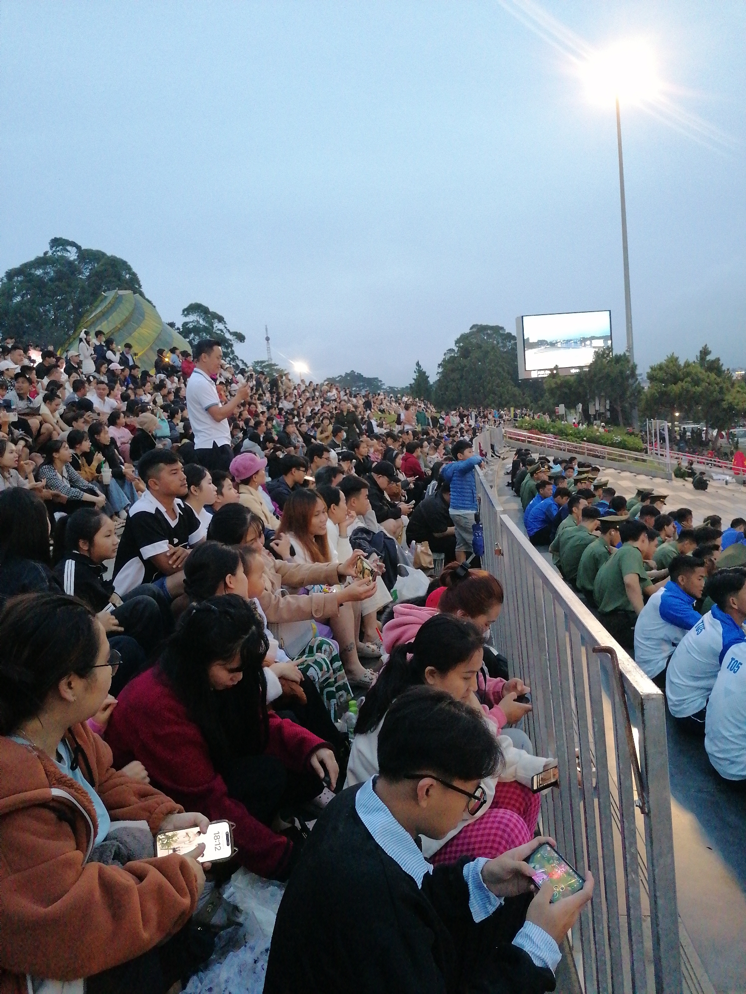 The image depicts a large crowd of people seated on tiered stadium seating, appearing to be attending an outdoor event at dusk. The setting is the Da Lat Stadium in Da Lat, Vietnam, identifiable by the stadium's architecture and the surrounding natural landscape. In the background, a large screen displays an image, likely for the audience's viewing. A bright stadium light is also visible, illuminating the area. Many individuals are looking towards the screen or engaging with their mobile phones. The crowd consists of people of various ages, dressed in casual attire. Some individuals are actively holding and using their phones, possibly to take photos or videos of the event. The overall atmosphere suggests a communal gathering, possibly a sporting event or a concert, taking place in the late afternoon or early evening.
