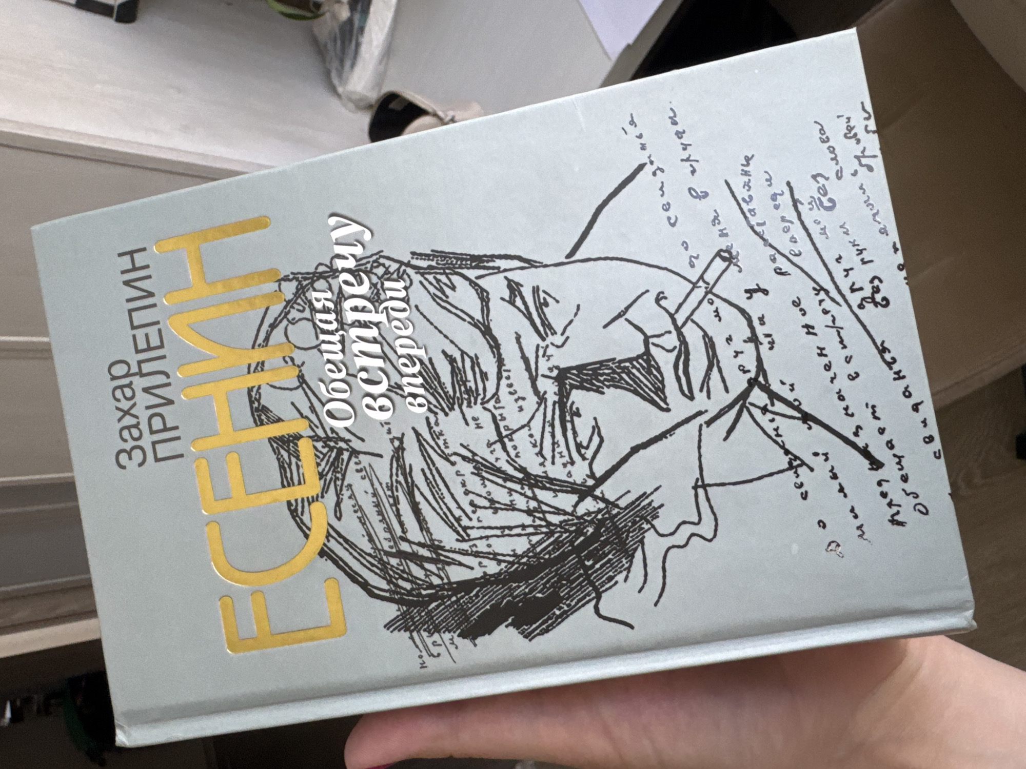 Here is a detailed description of the image:
The image shows a close-up of a book titled "Есенин. Обещая встречу впереди," by Zakhar Prilepin. The book has a light gray cover with the title and author's name printed in gold. The cover features a black and white sketch of Sergei Yesenin, the Russian poet, smoking a cigarette. There are also handwritten notes and fragments of text surrounding the portrait, giving it an artistic and literary feel. The book is being held in someone's hand.
The location is unknown, and the image does not provide specific details about Razdolye or any other city.