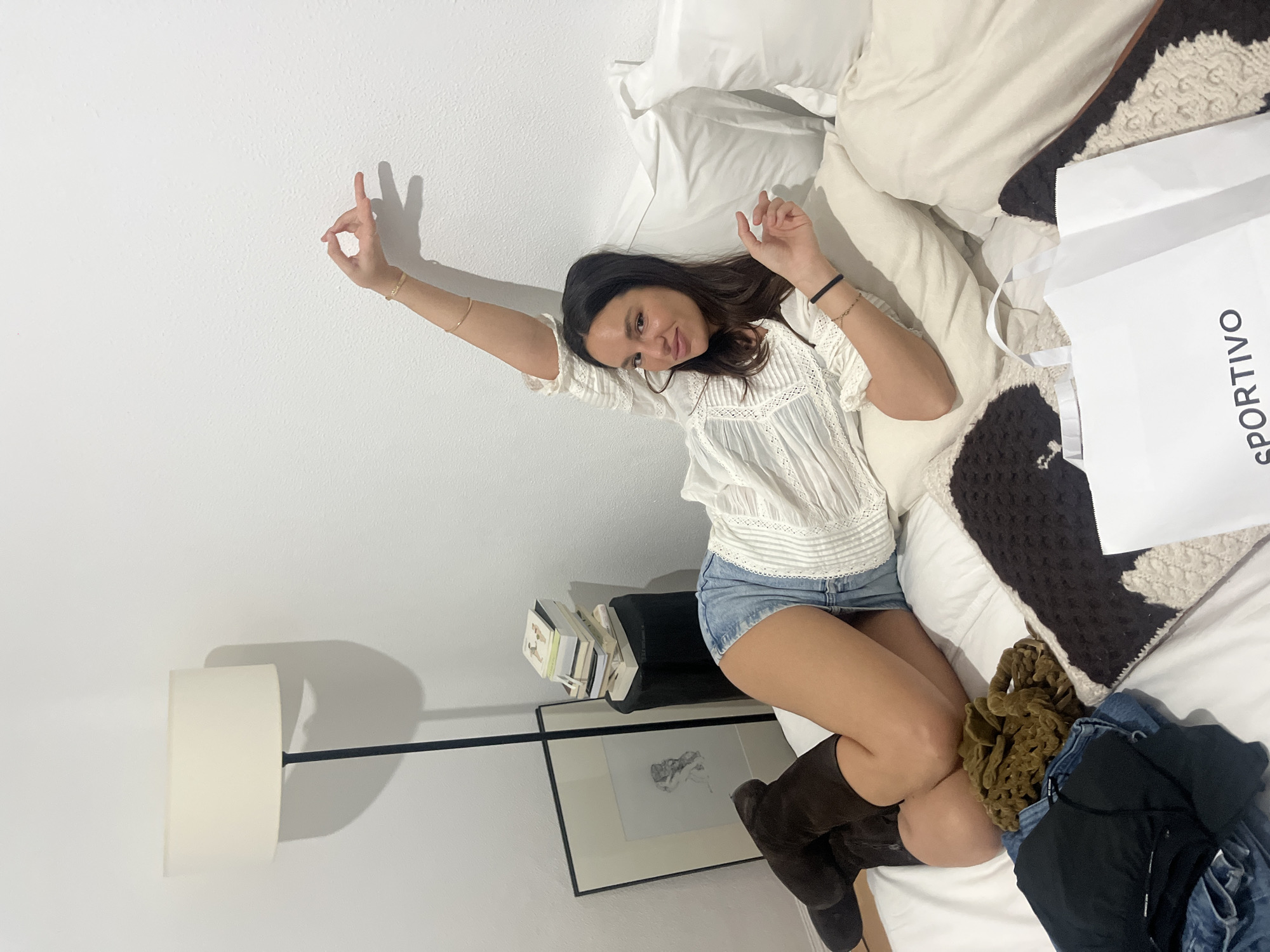 A woman is reclining on a bed with white linens and decorative cushions. She wears a white lace-trimmed blouse and denim shorts, complemented by dark brown boots. To her right is a white shopping bag with the word "SPORTIVO" printed on its side. Beside the bed stands a floor lamp with a white shade and a black base. A framed drawing rests on a ledge next to stacked books. The setting is a room with white walls. There is no information about the image's location.