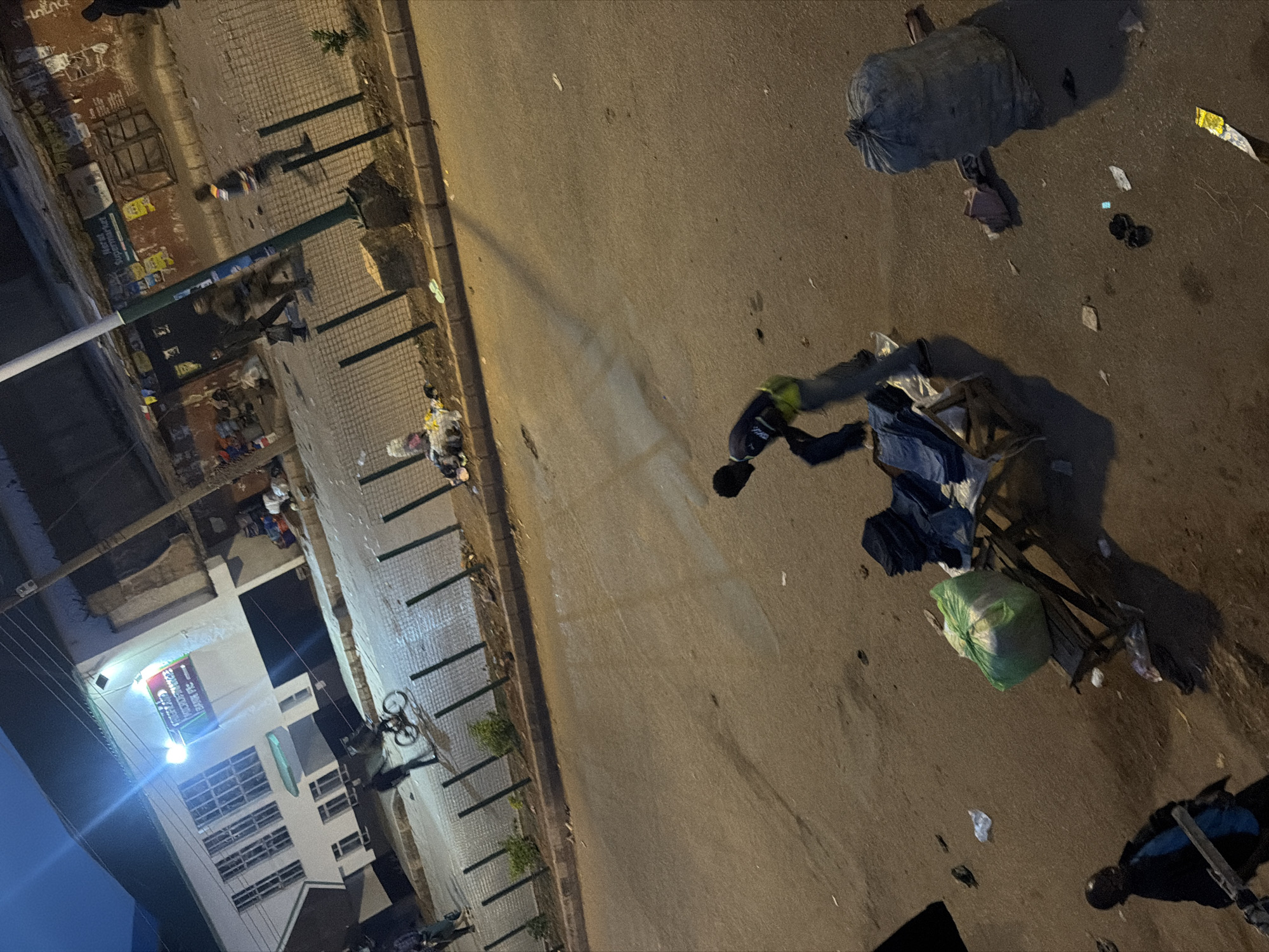 In Bukuru, Nigeria, at night, a street scene unfolds. A building, possibly a bank, with prominent signage, is illuminated. Across the street, a roadside market is visible, with various goods displayed on makeshift tables. Several individuals are present, some appearing to be vendors or shoppers. One person is seen interacting with a stack of clothing, possibly arranging it for sale or display. Another individual is walking with what appears to be a bicycle. The ground is littered with debris, including plastic bags and other trash, suggesting a busy and perhaps unkempt urban environment. A large, dark-colored trash bag sits on the asphalt. The lighting is a mix of streetlights and the illumination from the building, casting long shadows and creating a somewhat moody atmosphere.