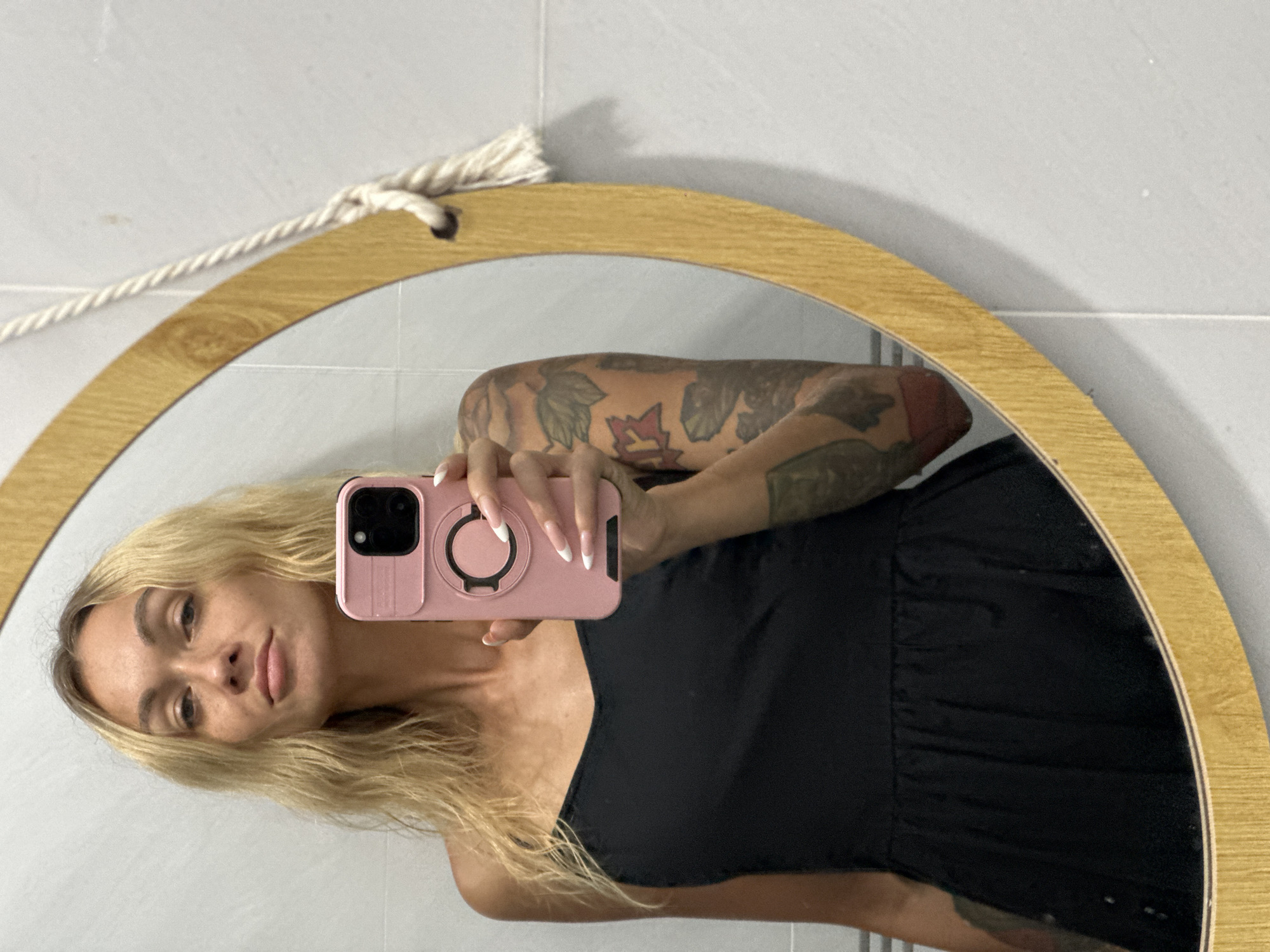 In Mũi Né, Vietnam, a young woman with blonde, wavy hair and freckles takes a mirror selfie. She is wearing a black dress and holding a pink phone in her right hand. Her left arm, visible in the reflection, is covered in tattoos. The mirror is round with a wooden frame and a rope hanger. The background appears to be a tiled wall.