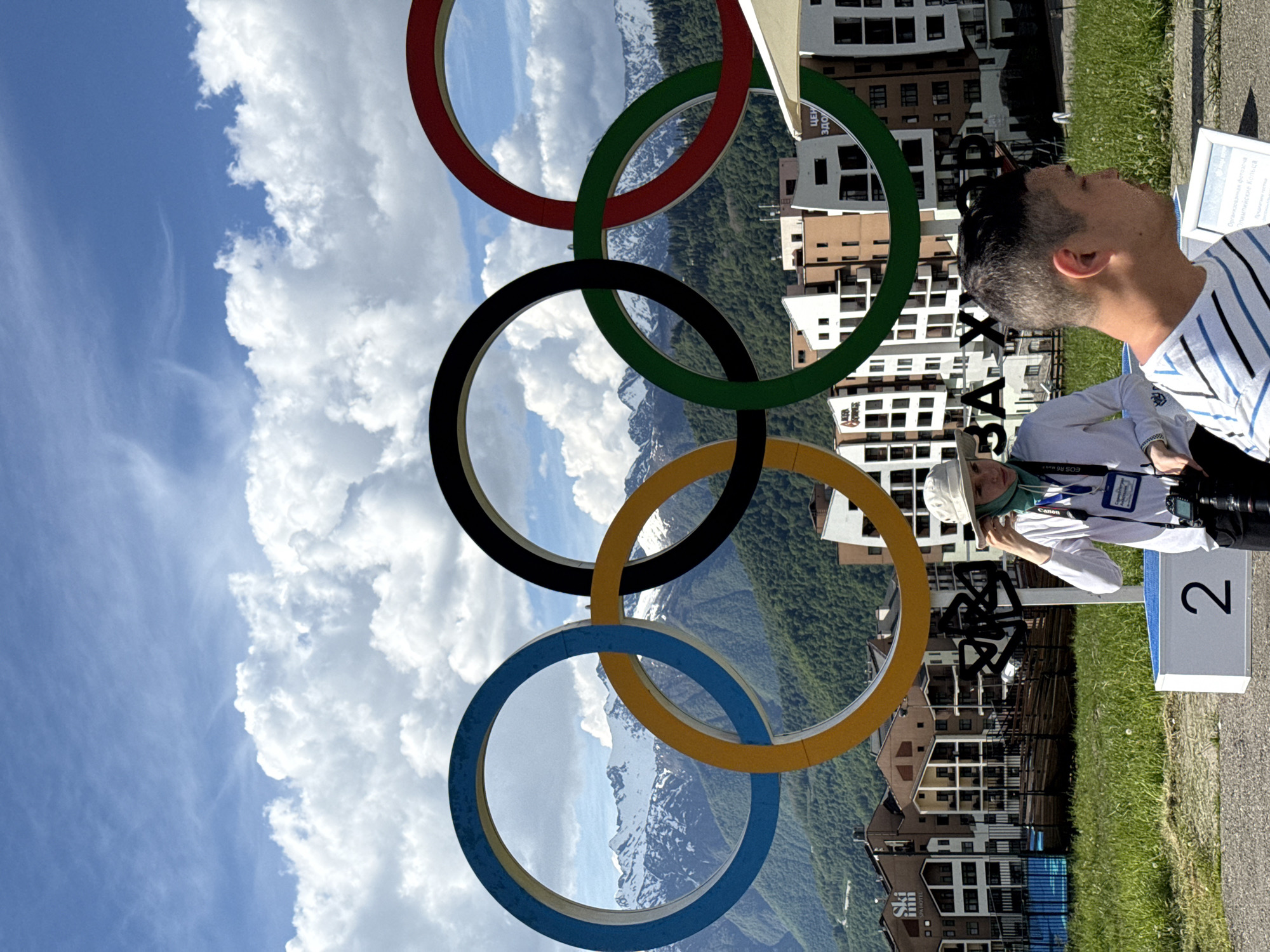 This media file captures a bright, sunny daytime scene, likely in a mountainous resort area, featuring a large Olympic rings sculpture. The image appears rotated 90 degrees clockwise from its intended orientation.
In the foreground, a man with short, dark hair, wearing a striped blue and white shirt, is visible from the chest up, looking towards the top right of the frame (upward when oriented correctly). To his left, slightly behind him, another person wearing a light-colored hat and shirt is holding a camera (a Canon EOS R6 Mark II is visible on the strap), seemingly preparing to take a photo or observing the surroundings. This person also wears an identification badge around their neck.
The prominent central feature is a massive Olympic rings sculpture, with the rings colored blue, yellow, black, green, and red from left to right (when upright). These rings frame a picturesque background of snow-capped mountains and lush green forests. Below the mountains and behind the rings, several multi-story buildings with modern architecture, resembling hotels or residential complexes, are visible. One building has "SOHO HOTEL" signage.
The ground in the mid-ground consists of green grass and a paved pathway. A white box with the number "2" is on the right side of the frame, and a small sign next to it has partially visible text "ОРГАНИЗАЦИОННАЯ ПРОГРАММА олимпийский команда" (Organizational program, Olympic team). Another sign, partially obscured by the Olympic rings, seems to read "ЦЕНТ ЗДО," possibly part of "Центр Здоровья" (Health Center). The sky above is blue with scattered fluffy white clouds. The setting, with the Olympic rings, mountains, and resort-like buildings, strongly suggests a former Winter Olympic Games venue, consistent with Estosadok, Russia.