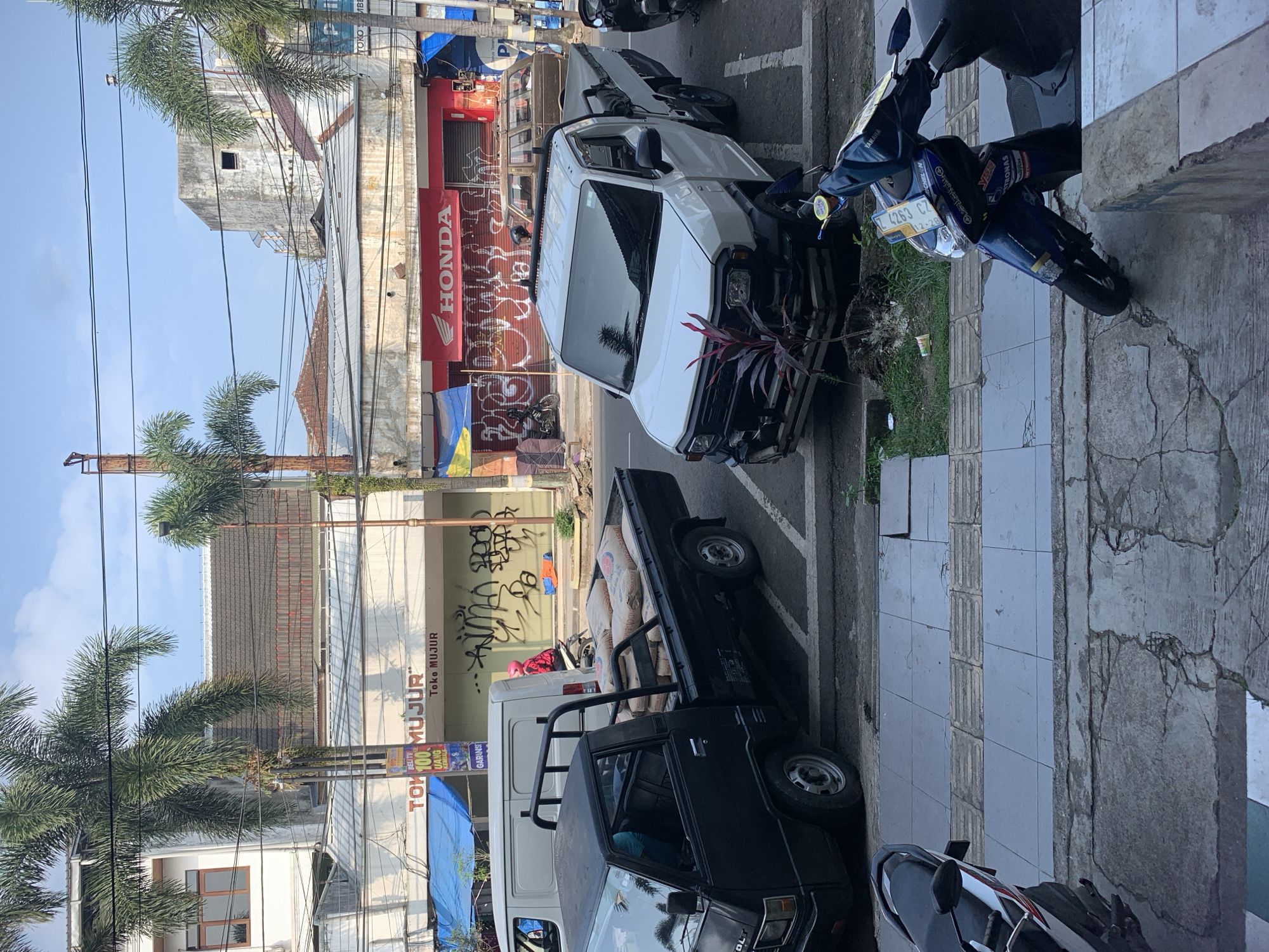 In Sumedang, Indonesia, a street scene unfolds under a bright sky, likely in the morning given the clear lighting. The foreground is dominated by a cracked concrete sidewalk and a section of grey tiled pavement. To the right, a blue and black Yamaha scooter is parked, angled towards the street, with a license plate reading "E 4263 CZ".
The street itself is paved and marked with white lines indicating parking spaces. A white pickup truck is parked in one of these spaces. To the left, a black pickup truck is also parked, its bed filled with numerous sacks of what appear to be cement or similar building material. A white van is parked behind the black pickup. A person wearing a pink jacket and a helmet is seated on a motorcycle to the left of the white van, looking to the right.
Behind the vehicles, a row of buildings lines the street. The buildings have awnings and storefronts. One storefront displays a large red "HONDA" logo. Another storefront has signs with "TOKO MUJUR" and "BELI TV 100 UANG KEMBALI". Graffiti covers the lower sections of some of these buildings. Palm trees are visible in the background, with their fronds reaching towards the sky. Power lines crisscross the scene overhead. The overall impression is one of an active commercial street in Indonesia.