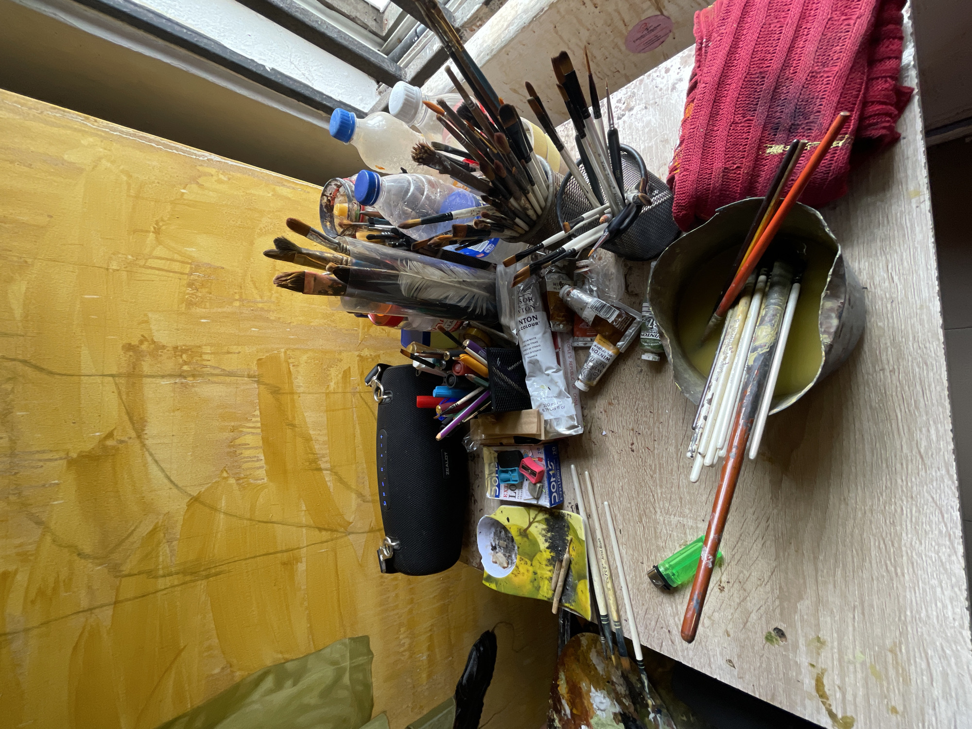 This is an overhead shot of an artist's workspace in Ado, Nigeria, featuring a collection of art supplies. The foreground is dominated by a variety of paintbrushes, some standing in a metal container filled with liquid, others lying scattered on a wooden table. Tubes of oil paint, including "WINTON OIL COLOUR," are also visible.
To the left, a large canvas painted in vibrant yellows and oranges is propped up. A black fabric case, labeled "ZEALOT," containing pens and pencils, hangs on the side. Several plastic bottles, some with blue caps, are also part of the setup. A bright red knitted cloth is draped over the supplies in the background.
The overall impression is one of creative activity, with supplies readily accessible. The lighting suggests it might be daytime, possibly indoors, given the clutter and arrangement of tools. The scene is unposed and candid, capturing the organized chaos of an artist's creative process. Visible text includes brand names like "ZEALOT," "WINTON," and "NTON," as well as product details like "200 ml" and "6.7508 fl oz".