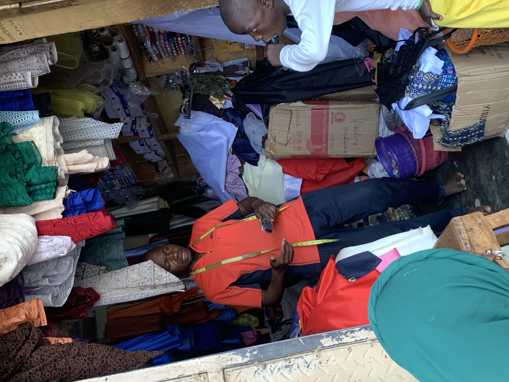 The image shows two men inside a cluttered space, possibly a fabric shop. One man in an orange shirt and dark pants lies among piles of colorful fabrics and holds a tape measure. The second man, wearing a white long-sleeved shirt, leans in close to the first man. The fabrics include lace and other materials in various colors. Packaging and other objects are scattered around the scene, along with an open cardboard box. OCR text suggests that the image was taken in Kafanchan, Nigeria.