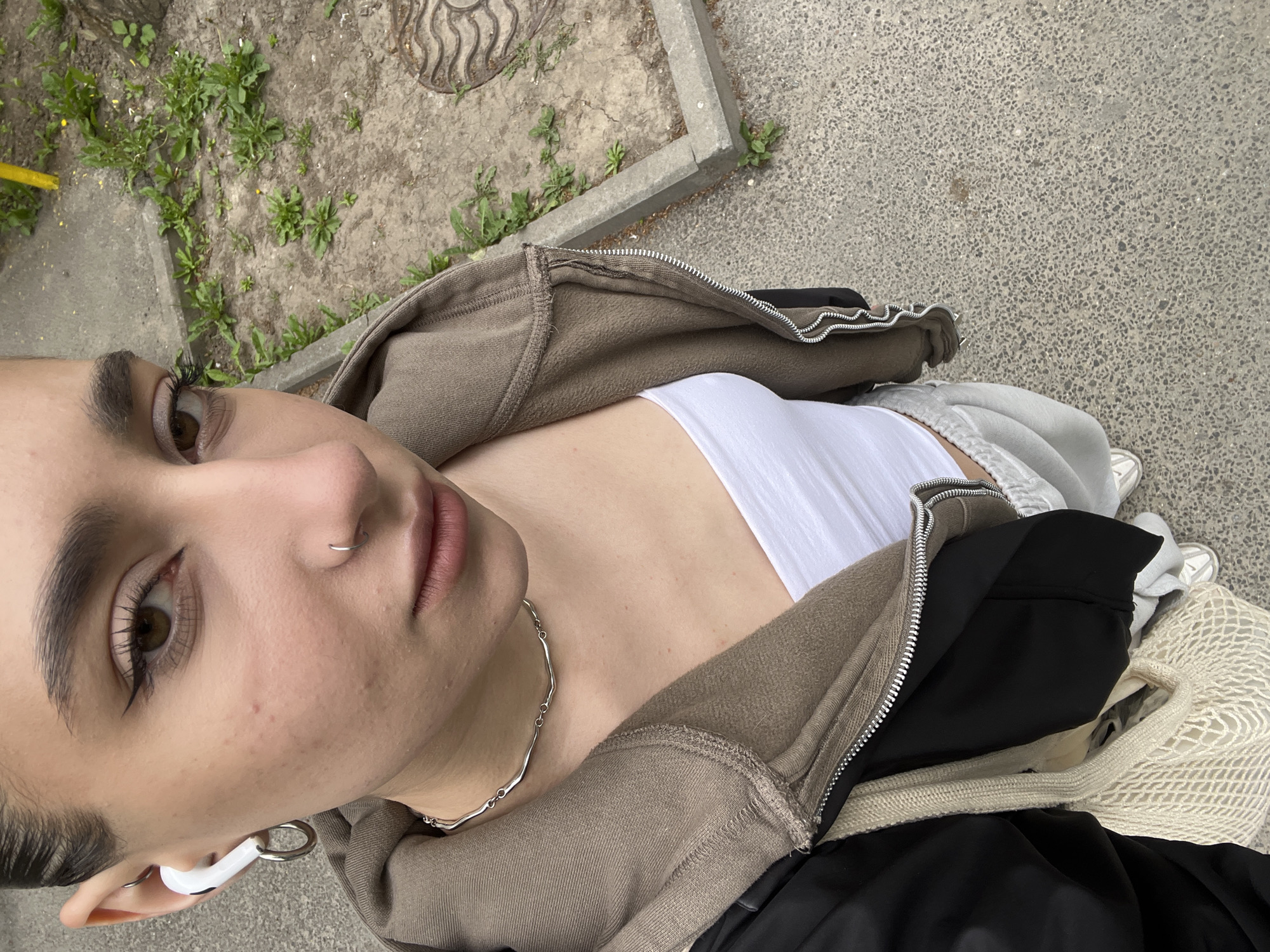 A person is photographed from a high angle outdoors. The individual has light skin, dark eyebrows, a nose piercing, and an earbud in the left ear. They are wearing a white top, a brown zip-up hoodie, and a silver necklace. A black garment and a net bag are also visible. The background shows a concrete surface with some vegetation, possibly near a sidewalk. Without additional evidence, I cannot confirm the location is Ternopil, Ukraine.