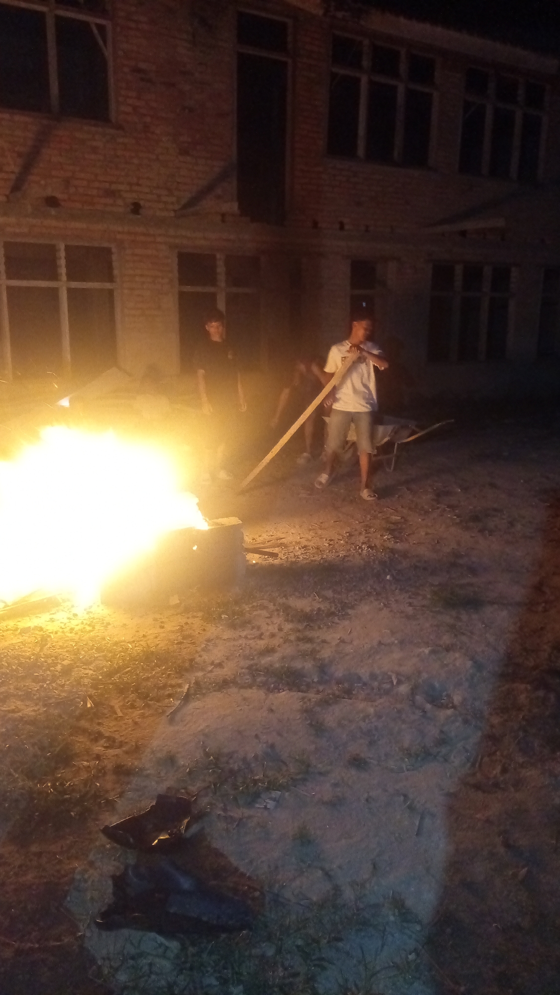 In the city of Unknown, Unknown, at night, two young men stand near a bonfire in front of a brick building with numerous windows. One man, wearing a black t-shirt, stands to the left of the fire. The other man, in a white t-shirt and shorts, is to the right of the fire, holding a long piece of wood. A wheelbarrow is visible behind him. The ground is dirt with sparse vegetation, and there is a shadowy object in the foreground. The building's windows are dark, suggesting it is unoccupied or unlit.
