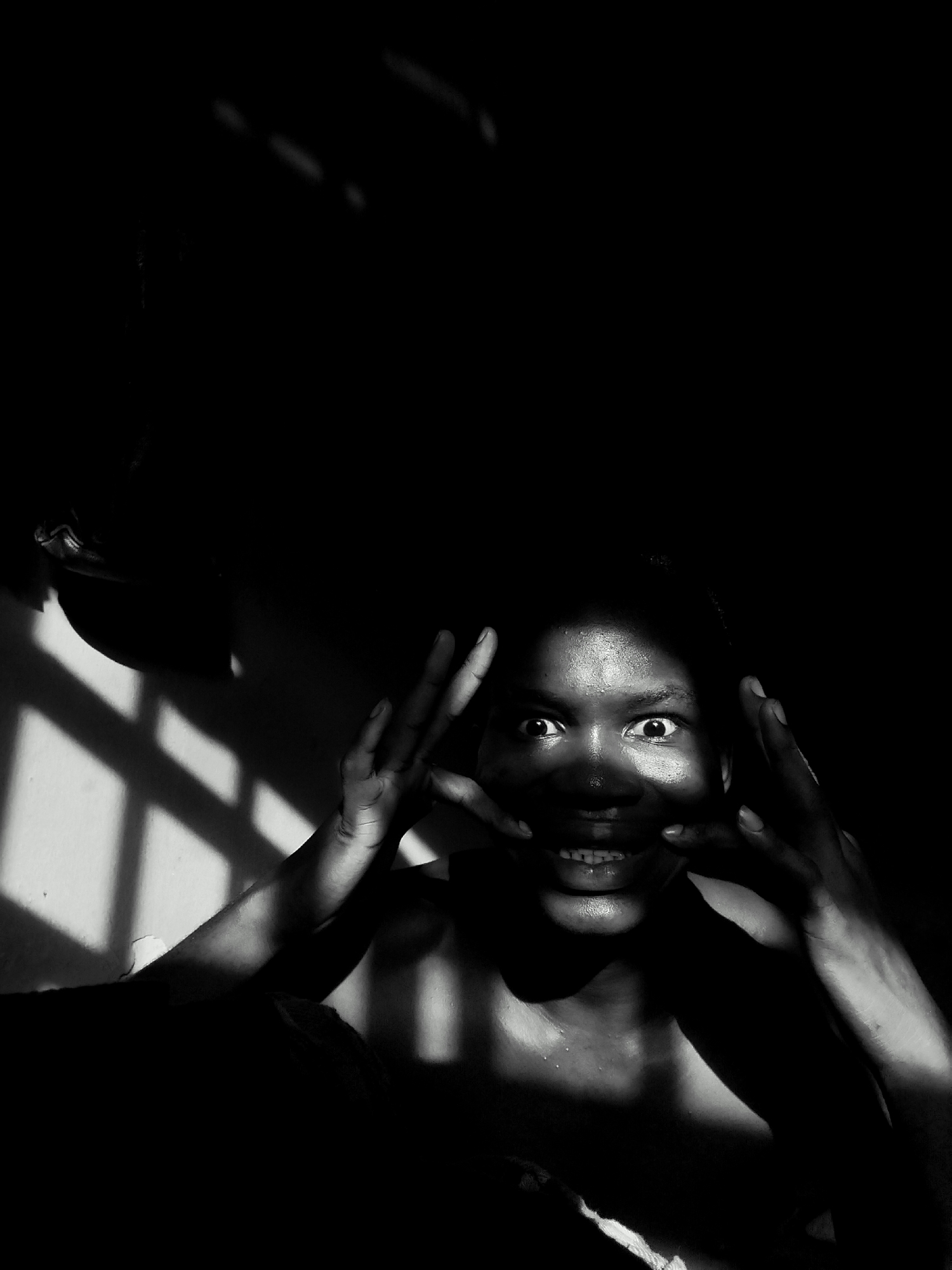 A monochrome, low-angle shot depicts a person illuminated by light filtering through a window, creating a stark contrast of light and shadow. The person's face is visible, with widened eyes and a grin revealing teeth. Their hands are raised, framing their face. Water droplets are visible on their skin. The location, Keffi, Nigeria, is assumed given the prompt. The background is predominantly dark.