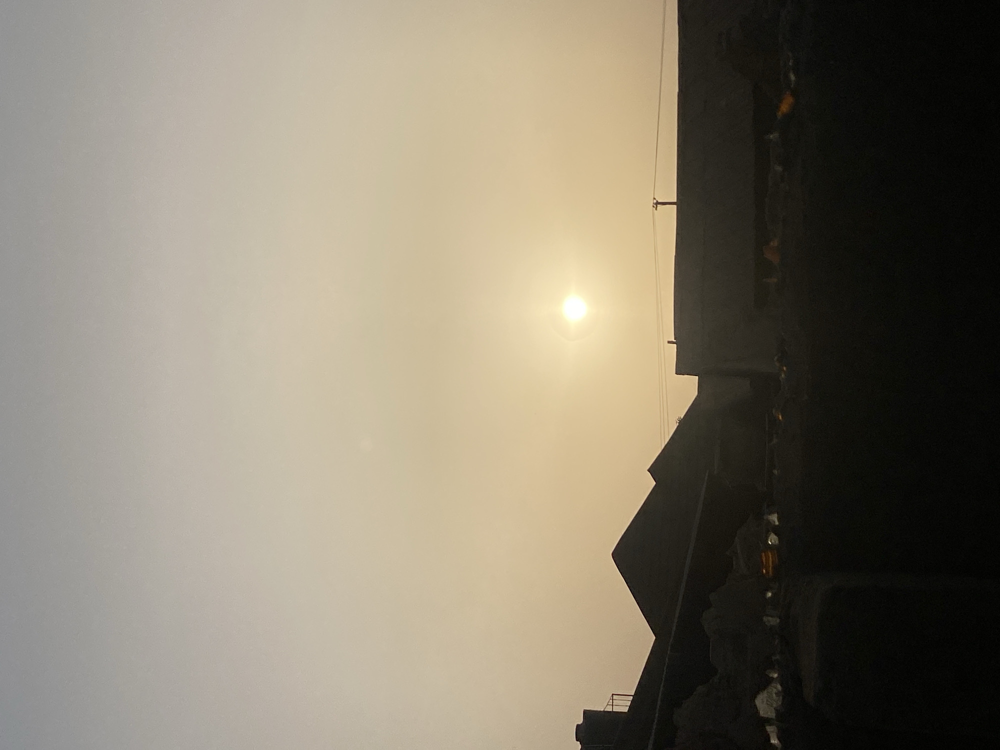 The image shows a bright sun in a hazy, light yellow sky. To the right are silhouettes of buildings. The photograph was taken in Narayi, Nigeria.