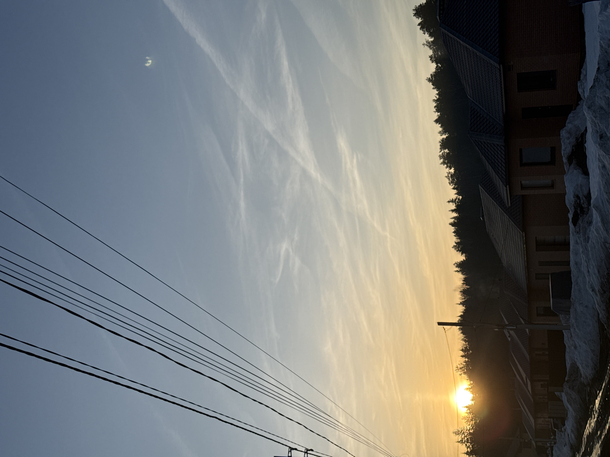 The image shows a sunrise in Chaikovsky, Russia. Power lines are visible in the foreground against a pale blue sky with contrails. The sun is rising behind a line of trees and a building with a dark roof. A snowbank is present on the right side of the frame.