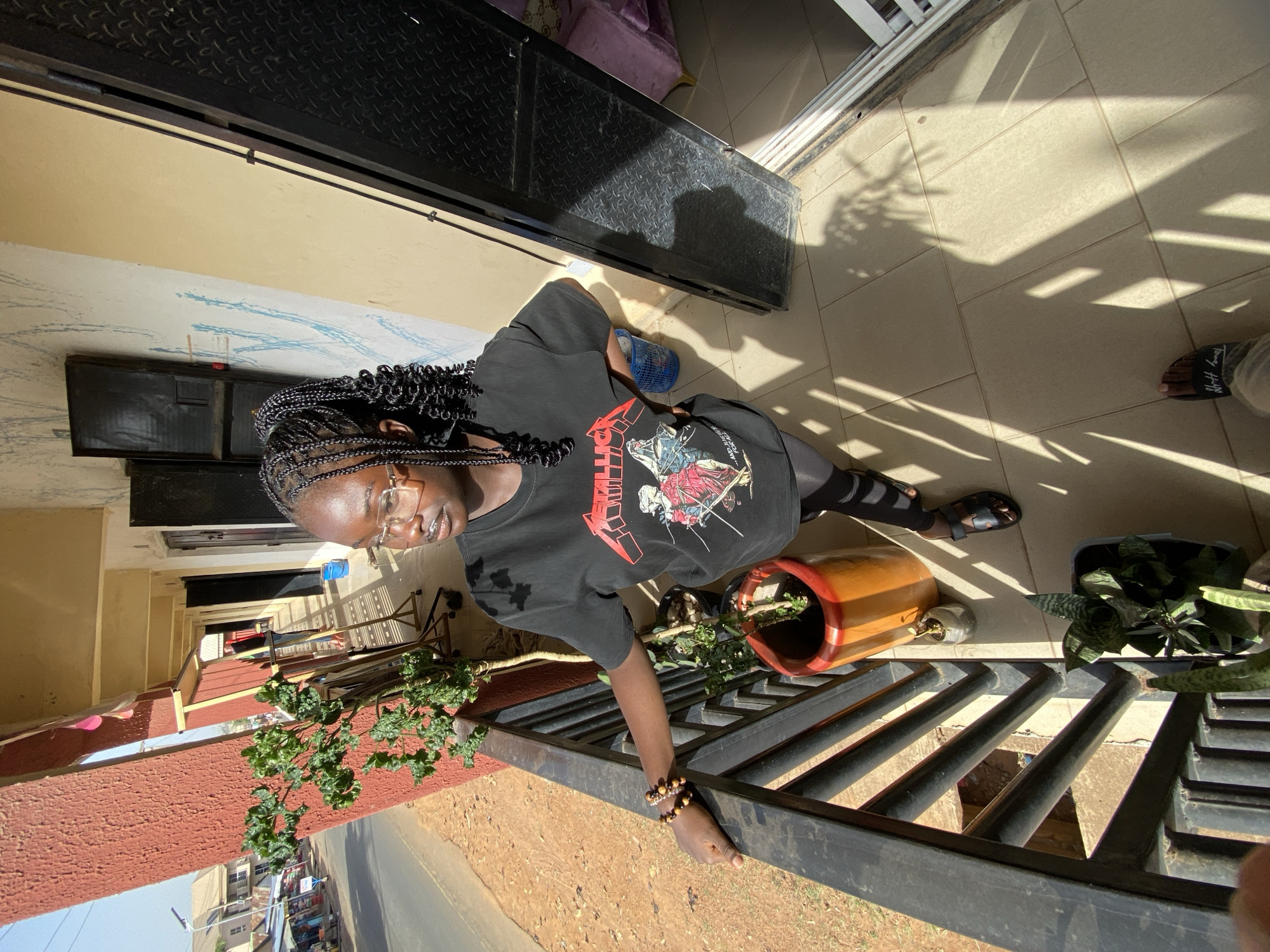 A young person with dark hair braided and wearing glasses is standing on a tiled patio or porch adjacent to a brown brick building. The person is wearing a black t-shirt with a graphic design and black leggings or tights. They are holding onto a black metal railing.
On the patio are potted plants and other objects. The foreground view shows an asphalt road, and across the road are buildings.
The location is determined to be in Bukuru, Nigeria.