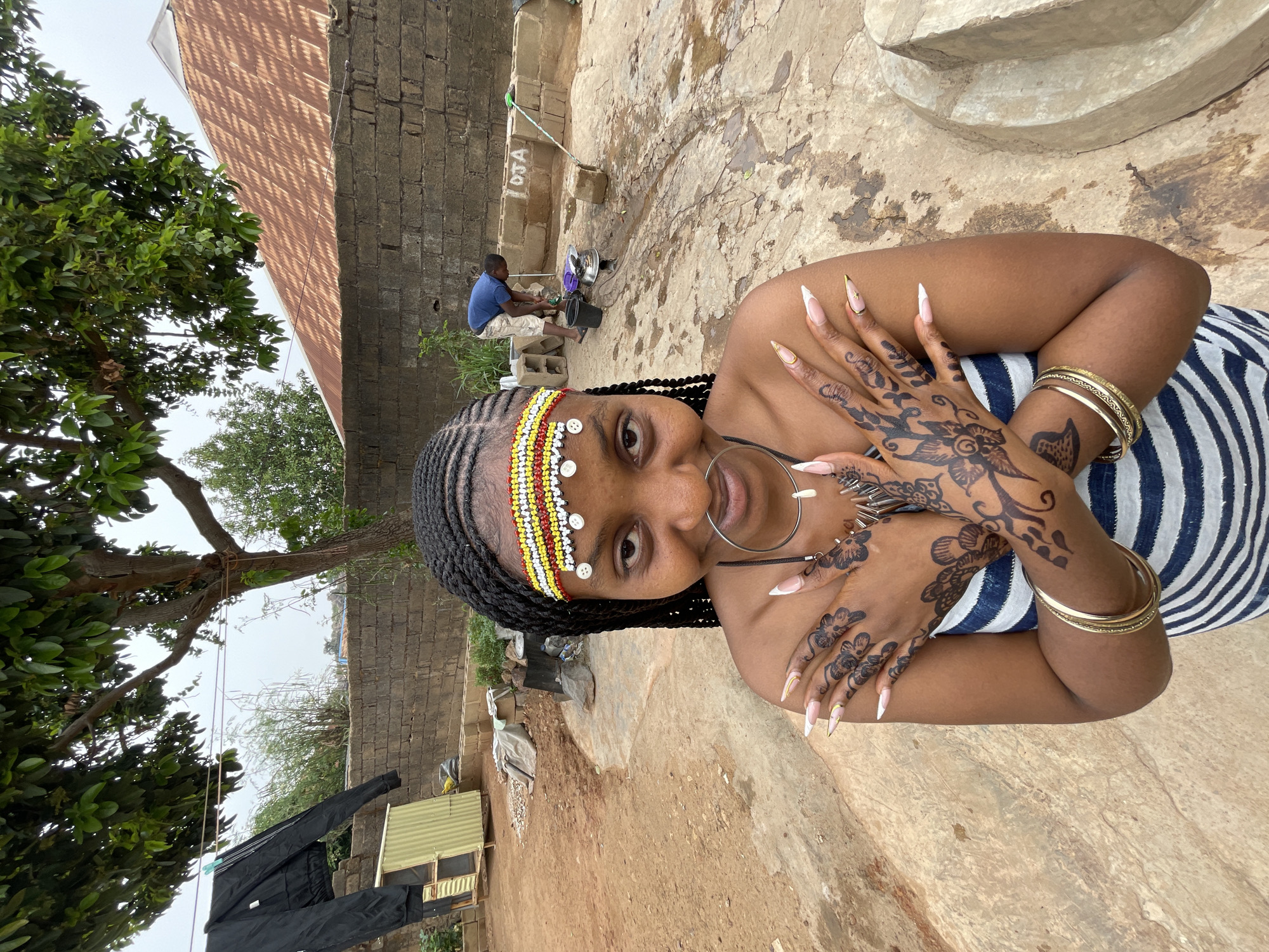 A young woman is the central subject of this outdoor photograph. She is adorned with a colorful beaded headband and has a large ring through her nose, and her hands are decorated with henna designs. She wears several gold bracelets on her wrists. She is wearing a striped top and has her arms crossed.
The background reveals a rural environment. A brick building with a metal roof stands behind her. A man is seen washing dishes outside near the building. Clothes hang on a line strung between trees. The ground is bare and appears to be concrete. The location is likely in the city of Jos, Nigeria.