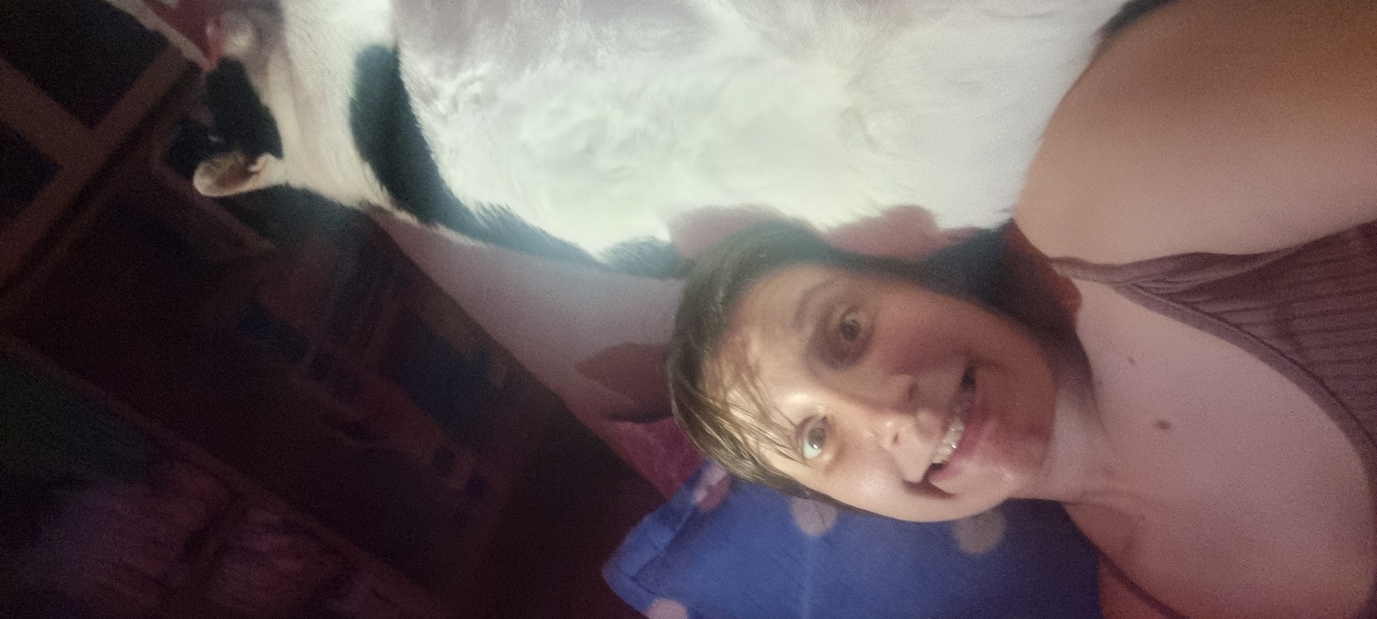 A selfie taken indoors depicts a woman with fair skin, short brown hair, and braces, smiling slightly at the camera. She wears a brown tank top. Above her is a white and black cat. A blue pillow and indistinct furniture are visible in the background. There is no verifiable indication of the location being Dunnae-myeon, South Korea.