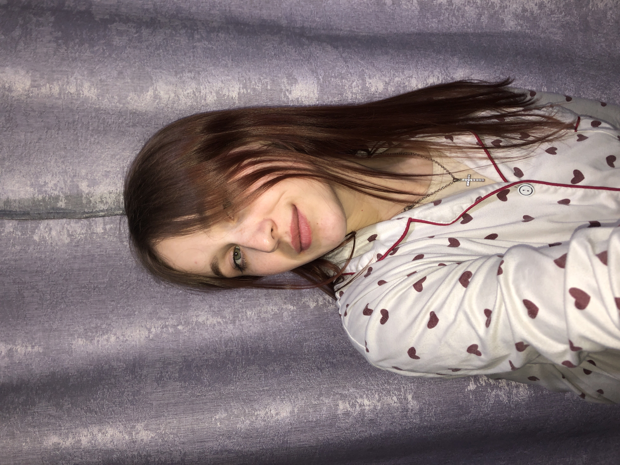 A young woman with light skin, long auburn hair covering part of her face, and green eyes is lying on a textured, grey surface. She is wearing white pajamas with small burgundy hearts and red piping, and a silver necklace with a cross pendant. The location is potentially within a room or building in Фоки, Russia, based on the user's prior query context. The scene appears to be indoors, possibly on a bed or couch. The lighting is dim, suggesting it could be evening or a low-lit room.