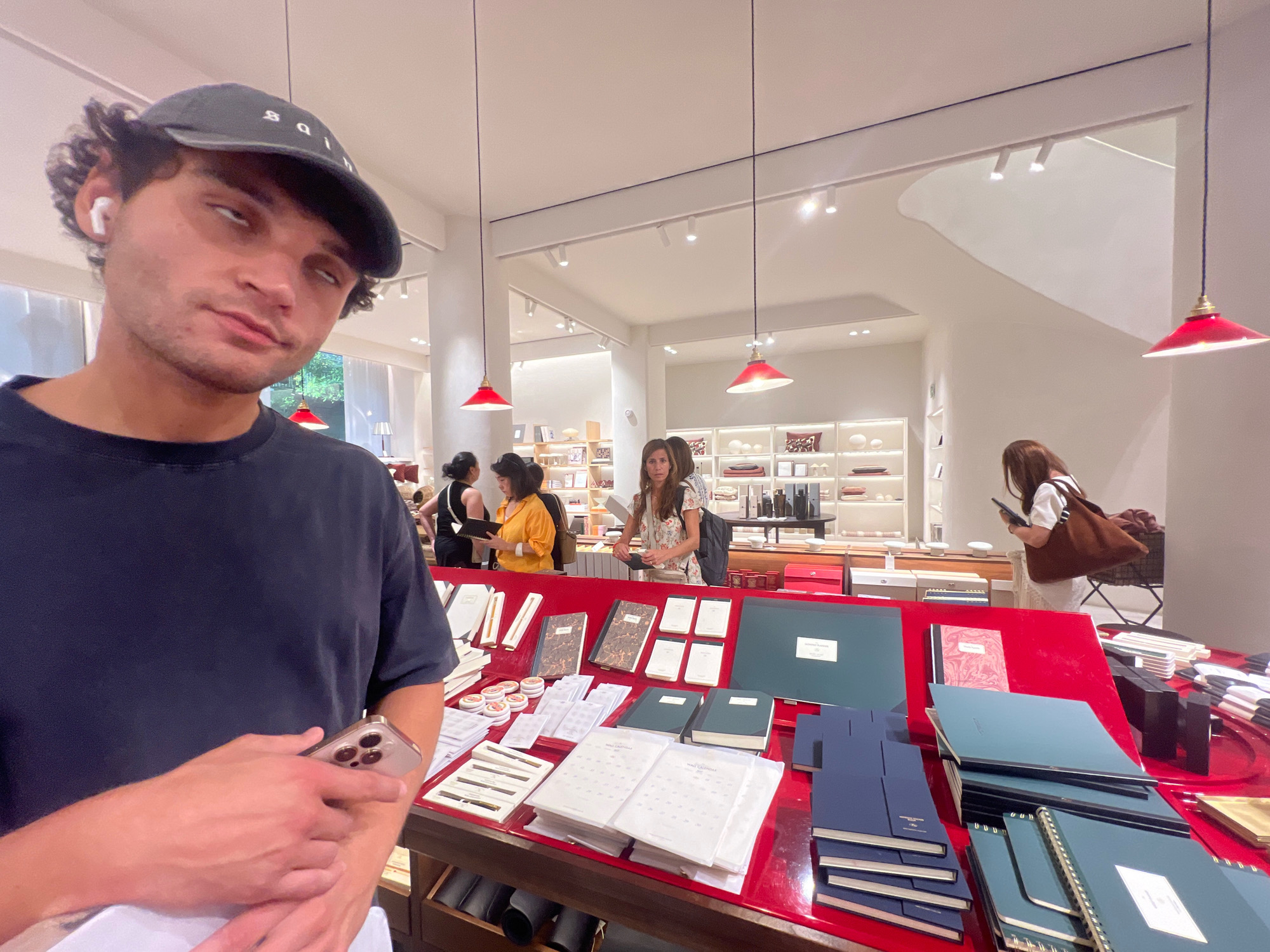 A young man wearing a dark t-shirt and a black baseball cap is in the foreground, looking towards the camera with his eyes rolled back. He is holding a smartphone in his right hand, which is positioned near his chest. The background shows a well-lit retail store, possibly a stationery or gift shop, with shelves stocked with various items like notebooks, boxes, and decorative objects. Several other people are visible in the store, browsing the merchandise or interacting with staff. Red lamps hang from the ceiling, and the overall ambiance suggests a modern and organized commercial space. The store appears to be located in Madrid, Spain, given the context provided.
