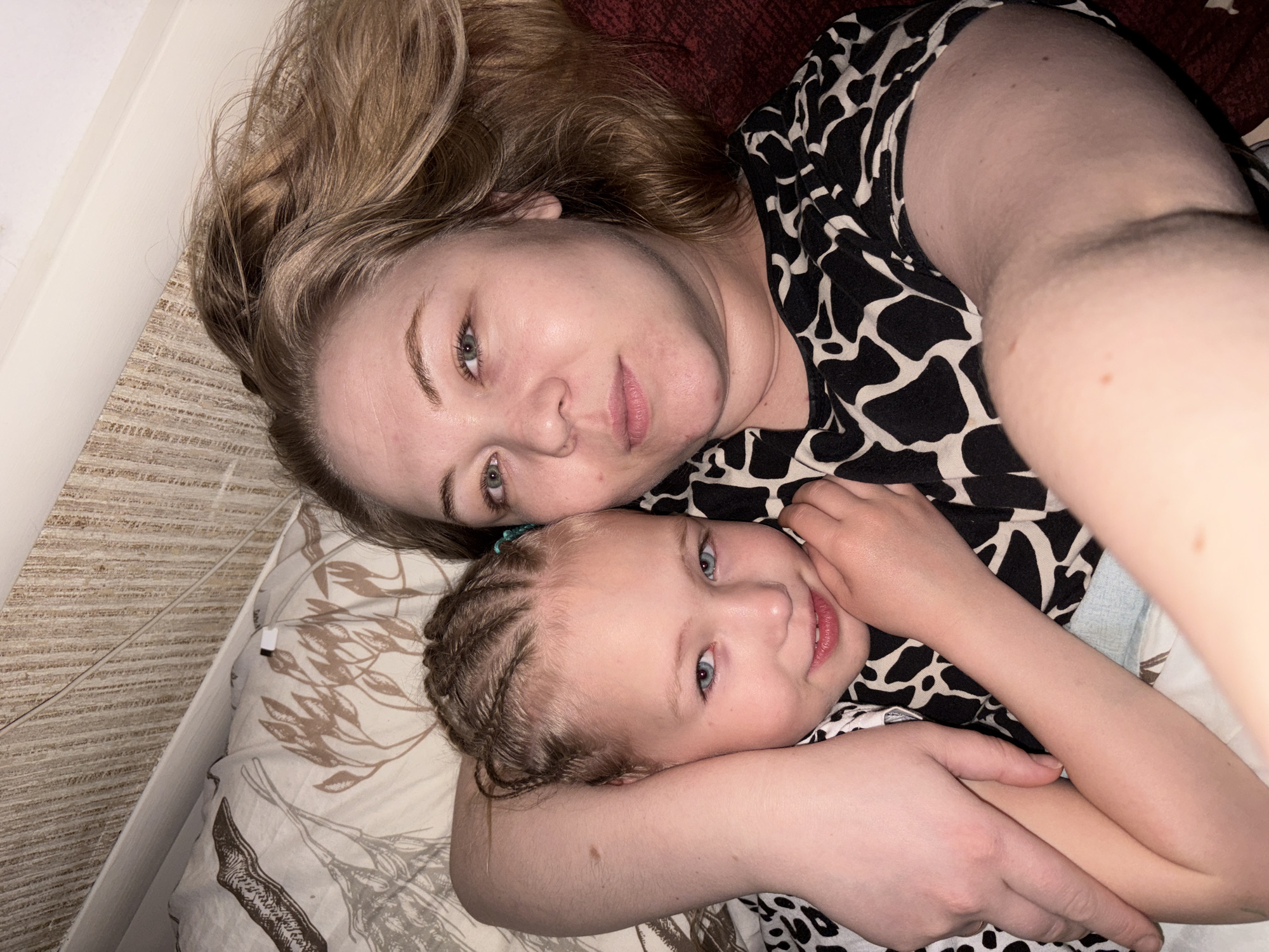 A woman and a young child are lying down on what appears to be a bed or similar surface. The woman is lying on her back with her head resting on a pillow. She has blond hair and is wearing a black and white patterned top. The child is lying on her side, with her head resting on the woman's arm. The child has light hair braided and is wearing a similar patterned top. The background is slightly blurred, but it appears to be a room with neutral-colored walls. The lighting is soft and diffused. There is no information related to the city of Noviy, Russia in this image.