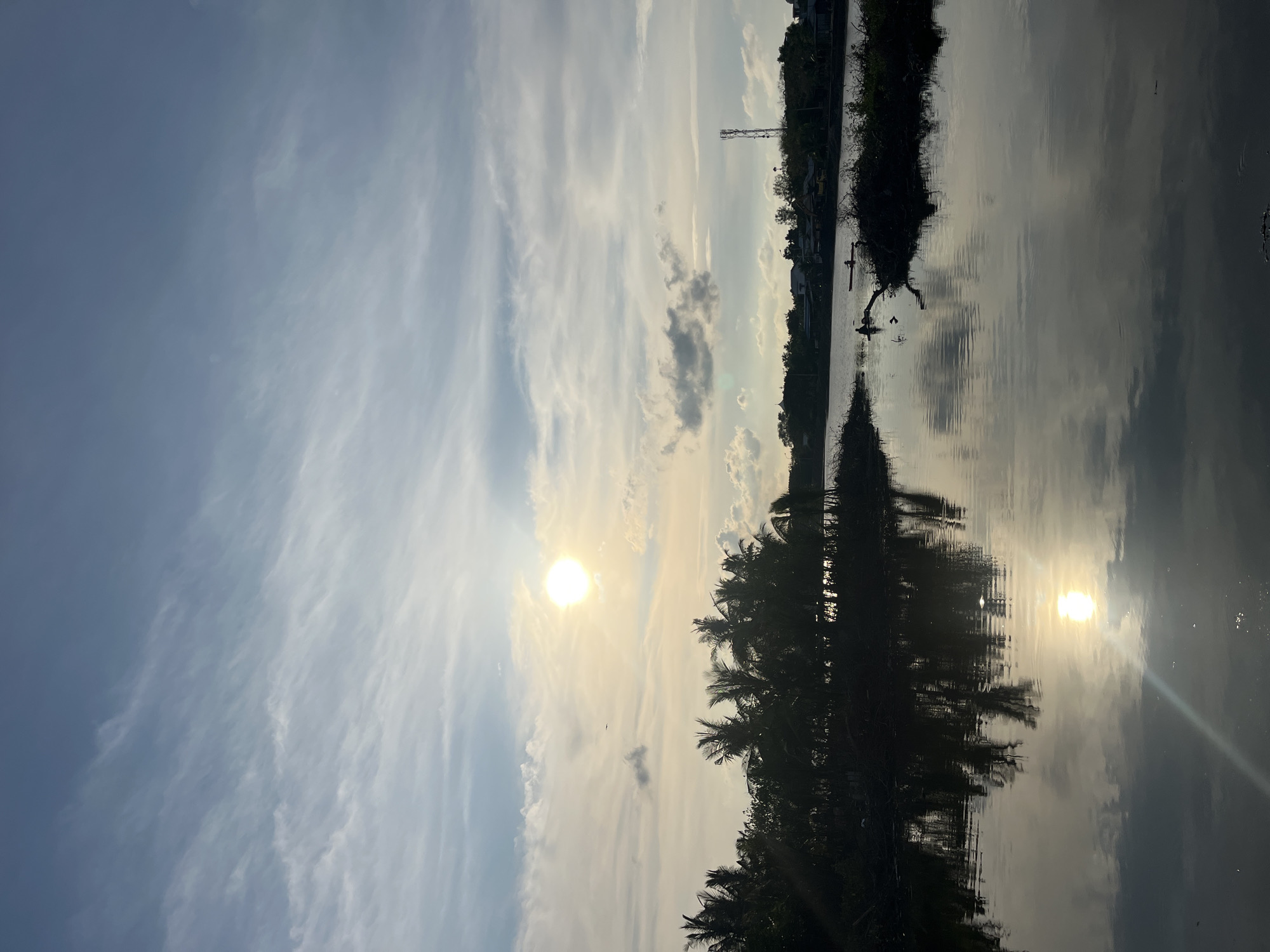 The image shows a waterfront scene, likely at sunset or sunrise, in or near Lhokseumawe, Indonesia. The sky has a mix of blue and light, wispy clouds. The sun is visible through the clouds, appearing as a bright circle. The water reflects the sun and the sky, creating a mirror effect. Trees and vegetation line the opposite bank of the water. A communication tower is barely visible on the far shoreline. The image is taken at an angle.
