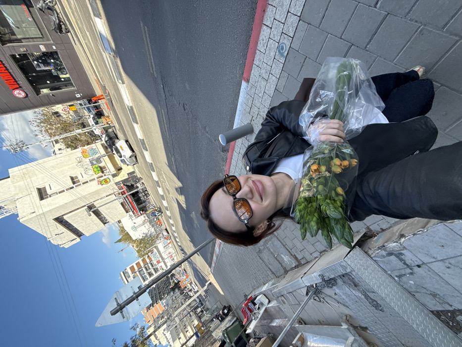 The image displays a female individual taking a selfie on Allenby Street in Tel Aviv, Israel, under a bright, clear blue sky. The woman, positioned in the foreground and facing the camera, has short, reddish-brown hair and wears dark-framed sunglasses with orange-tinted lenses. She is dressed in a black leather jacket over a white t-shirt and dark trousers, and holds a bouquet of green foliage and small orange, flower-like elements, encased in clear plastic, in her left hand. She exhibits a slight smile.
She stands on a light-grey paved sidewalk. To her immediate left, a green public waste bin and a metal ramp or grating are visible. To her right, a dark grey bollard stands near the curb. The street is asphalt, featuring a white and black striped pedestrian crossing and a red and white painted curb. Several vehicles are parked along the street, including a white Peugeot 208. In the midground and background, multiple urban buildings line the street. Prominently featured on the left side of the street is a distinctive light-colored, multi-story building with geometric architectural elements and visible green and yellow graffiti tags, identifiable as Beit Ha'Amudim (Pillars House). Further right, another building houses a Yamaha dealership, with motorcycles visible through its glass storefront. Additional buildings and trees are present further down the street.