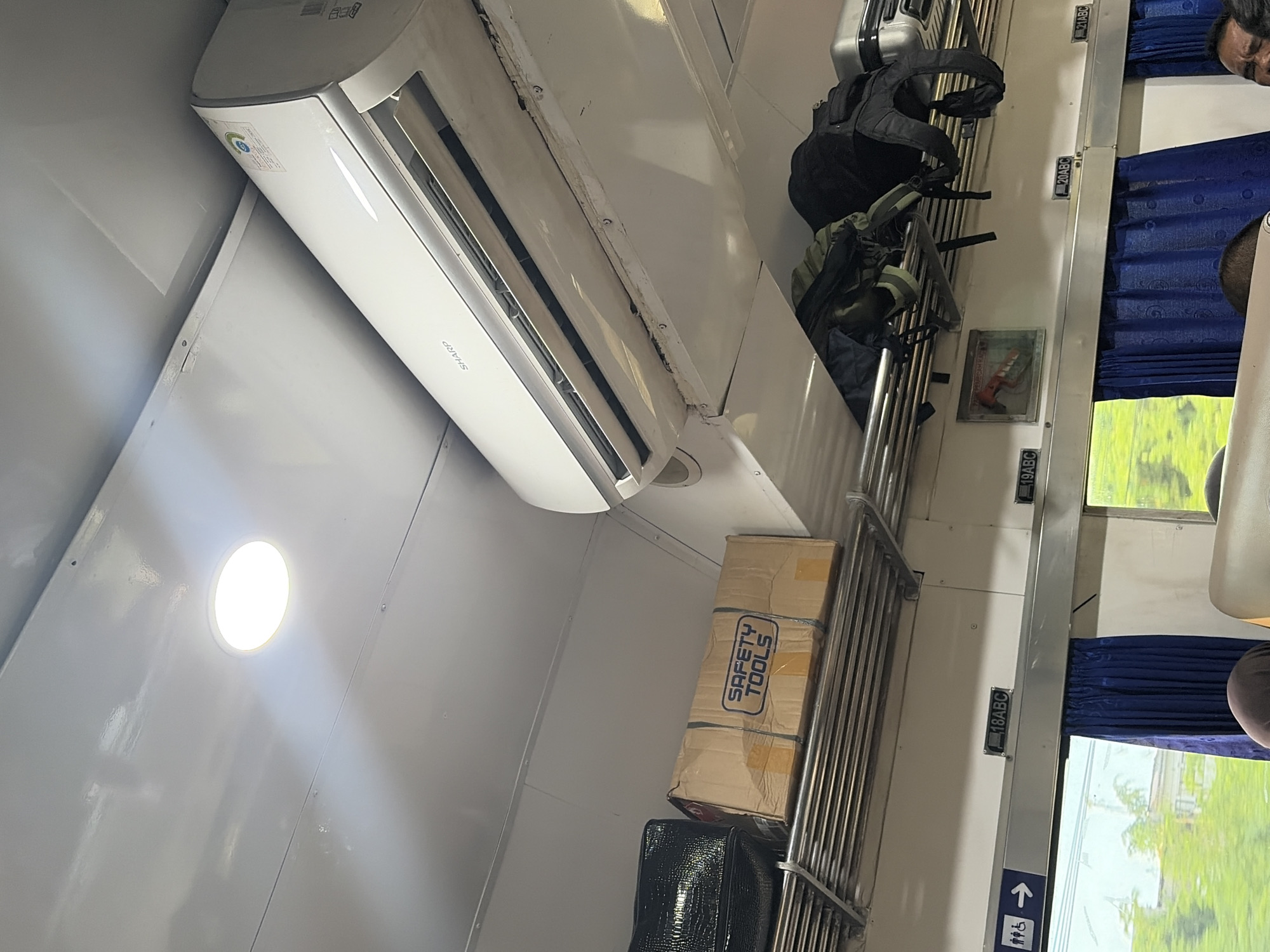The image depicts the interior of a train car, likely in Pekalongan, Indonesia, based on the context provided. The ceiling of the train features a circular light fixture and a white Sharp air conditioning unit. Below the AC unit, on a metal rack, are two black bags and a cardboard box labeled "SAFETY TOOLS". Further down, another metal rack holds several backpacks. A sign with an accessibility symbol and an arrow is visible on the wall, along with numbered signs like "18ABC". Blue curtains obscure a window, through which a blurred green landscape can be seen. The overall impression is one of functional, albeit somewhat worn, interior of a train carriage with luggage stowed overhead.