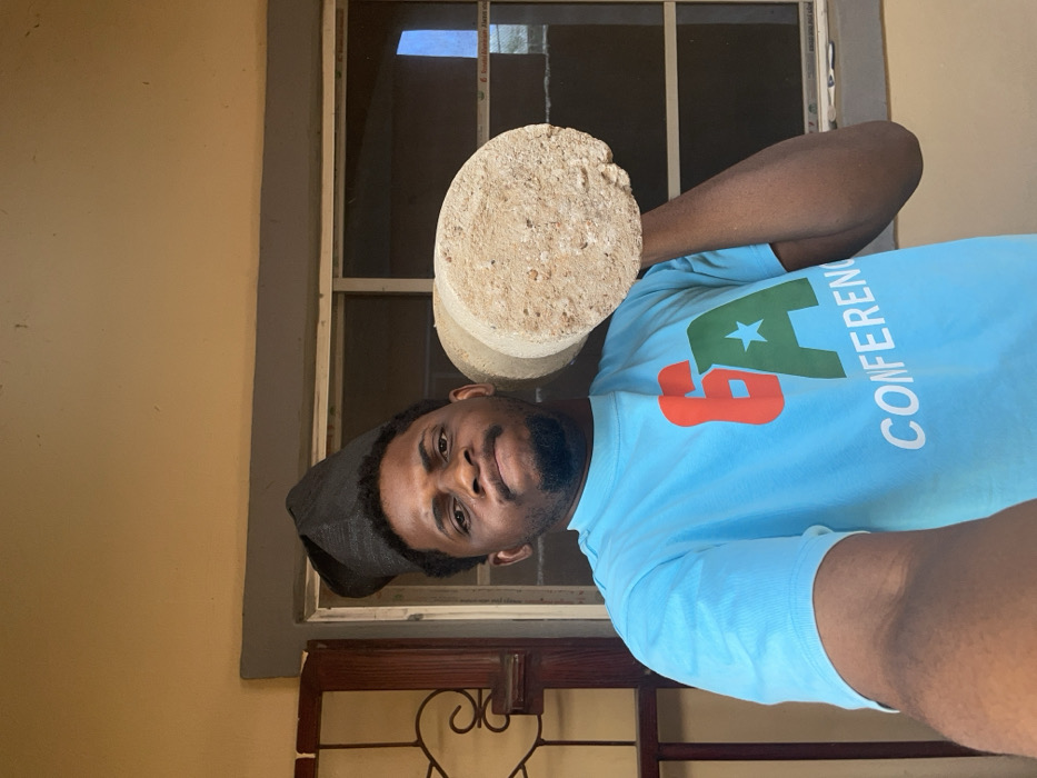 A male individual, presumed to be in Ube, Nigeria, is depicted in a chest-up shot. He has a dark complexion, short dark hair, and a trimmed beard, wearing a black, patterned traditional cap. His facial expression is firm, with direct eye contact towards the camera. He is clad in a light blue short-sleeved t-shirt displaying a logo featuring a red number '6', a green letter 'A' with a white star, and the word "CONFERENCE" in white text below. The individual holds a light beige, rough-textured cylindrical object, consistent with a concrete test cylinder, on his left shoulder, supported by his left arm. The background consists of a light beige wall. To the left, a portion of a dark brown wooden door frame with carved details is visible. Centrally located behind the individual is a window with visible horizontal white or light-colored frame divisions, possibly security bars, and a thin, metallic-colored frame. The lighting indicates a daytime setting.