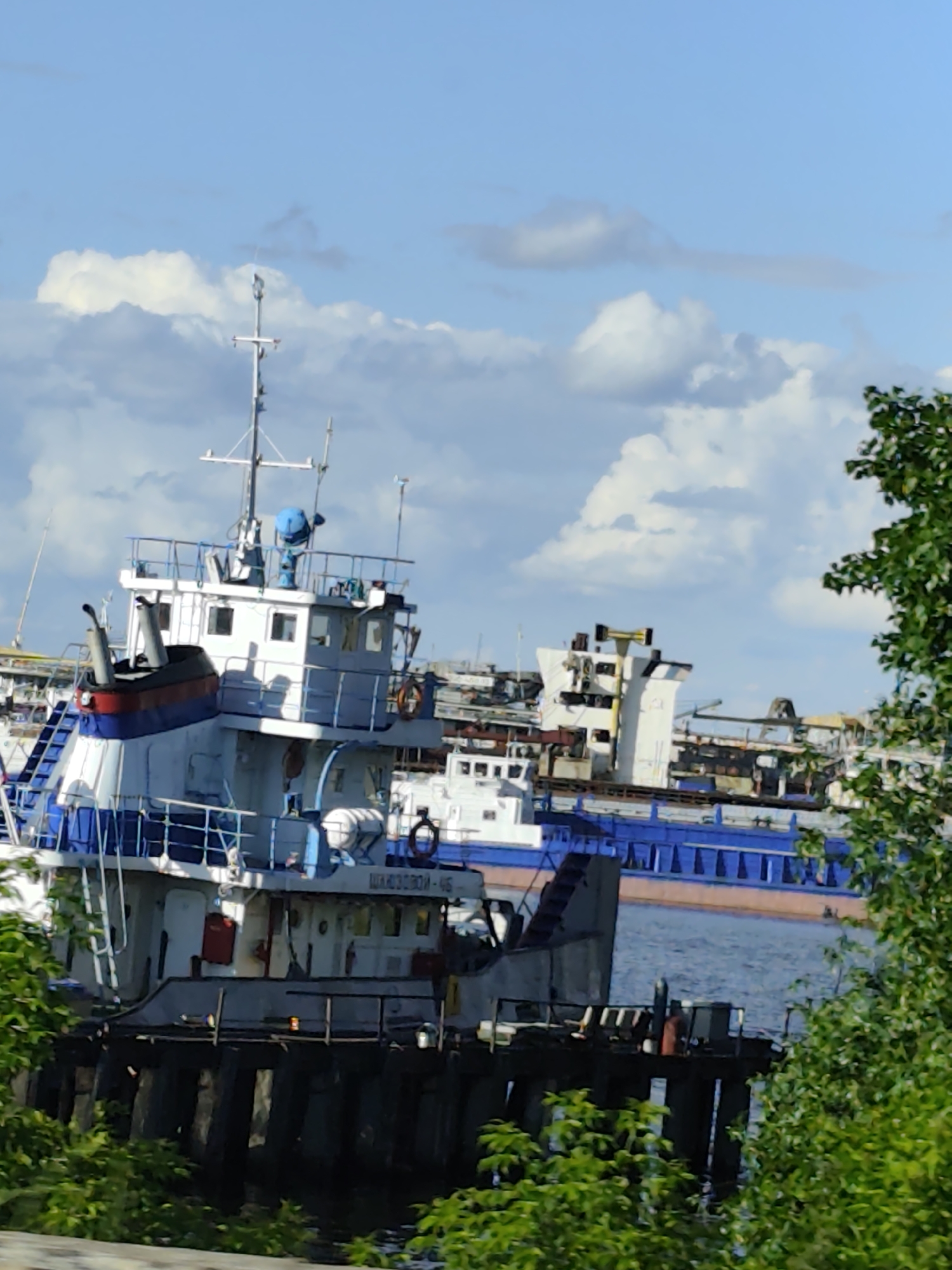 This image captures a section of the Kama River in Chaikovsky, Russia, featuring several vessels docked along the shore. In the foreground, a white and blue tugboat is prominently positioned, its superstructure adorned with railings and a prominent antenna mast. To its right and slightly behind, another vessel, larger and more industrial in appearance with a white superstructure and a blue hull, is visible. Further in the background, more industrial structures and vessels suggest a port or shipyard environment. The sky above is a clear blue, punctuated by scattered white clouds. Lush green foliage frames the lower left and right edges of the scene, hinting at the natural surroundings of the riverbank. The overall impression is that of an active waterway within an industrial area, under a bright, partly cloudy sky.