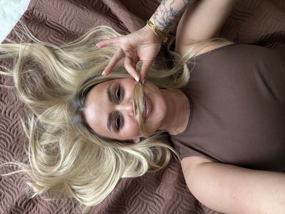 An overhead view depicts a blonde woman lying supine on a textured, brown quilted surface, identified as being in Sarapul, Russia. The woman, appearing in her 20s to 30s, is looking directly at the camera with a slight smile. She has light blonde hair, which is fanned out above her head. Her left hand, featuring neatly manicured white fingernails, is positioned near her face, holding a section of her hair just below her nose, creating the appearance of a mustache. She is wearing a plain, dark brown, sleeveless top. A gold-colored bracelet is visible on her left wrist, and a dark tattoo, partially obscured by her sleeve, is present on her left forearm. The background consists solely of the dark brown quilted fabric.