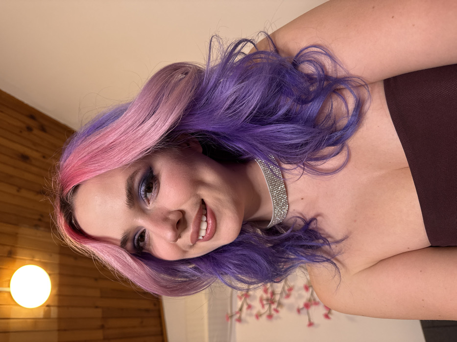A young adult female is depicted in a close-up, chest-up shot, smiling directly at the camera. She has shoulder-length, wavy hair dyed pink at the crown and purple on the lower sections. Her face features prominent blue eyeshadow, blush, and glossy lips, with dimples visible on her cheeks. She wears a strapless dark-colored top and a multi-row rhinestone choker necklace. In the background, a wooden slatted ceiling with a spherical white light fixture is visible in the upper left. A plain, light-colored wall occupies the right side, while a decorative branch with pink blossoms is partially visible on the left. The scene is located in the City of Zagreb, Croatia.