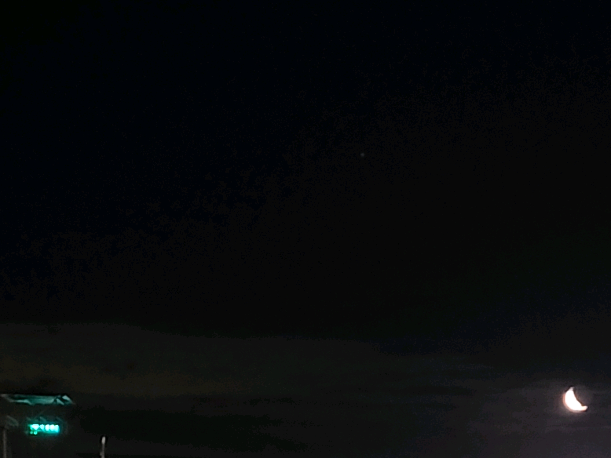 The image displays a dark night sky over Ozolnieki, Latvia. A crescent moon is visible in the lower right quadrant, illuminated by an unseen light source. In the lower left quadrant, a bright green light is emitted from what appears to be part of a traffic signal or illuminated sign. The rest of the scene is dark, with faint clouds or atmospheric haze present. No people are visible.