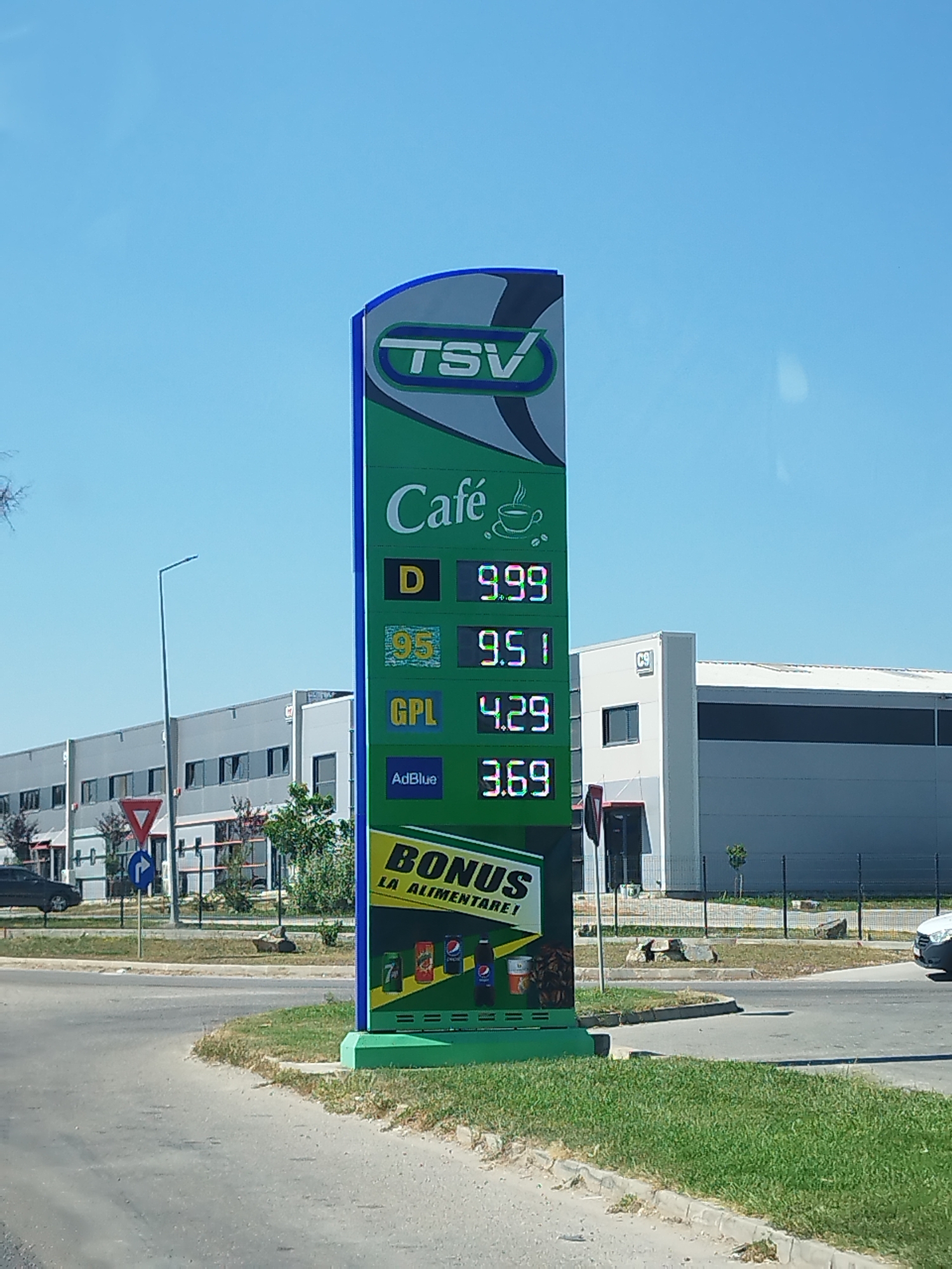 In the city of Buftea, Romania, a bright, sunny day is depicted with a clear blue sky. A prominent green and blue sign for TSV stands tall, displaying fuel prices and other offerings. The prices listed are 9.99 for "Café D", 9.51 for "95", 4.29 for "GPL", and 3.69 for "AdBlue". Below these, the word "BONUS" is prominently displayed in yellow with "LA ALIMENTARE!" written beneath it, suggesting a promotion related to food purchases. Images of various beverage cans are shown at the bottom of the sign.

In the background, a modern, light-colored industrial building complex with numerous windows is visible. A traffic light and a yield sign are positioned on the left side of the road, along with a white pole with a light fixture. Grass and some scattered rocks line the roadside. A dark-colored car is parked on the left, and a white van is seen on the right side of the road, suggesting ongoing activity. The scene captures a typical commercial area with clear visibility and a sense of everyday operation.