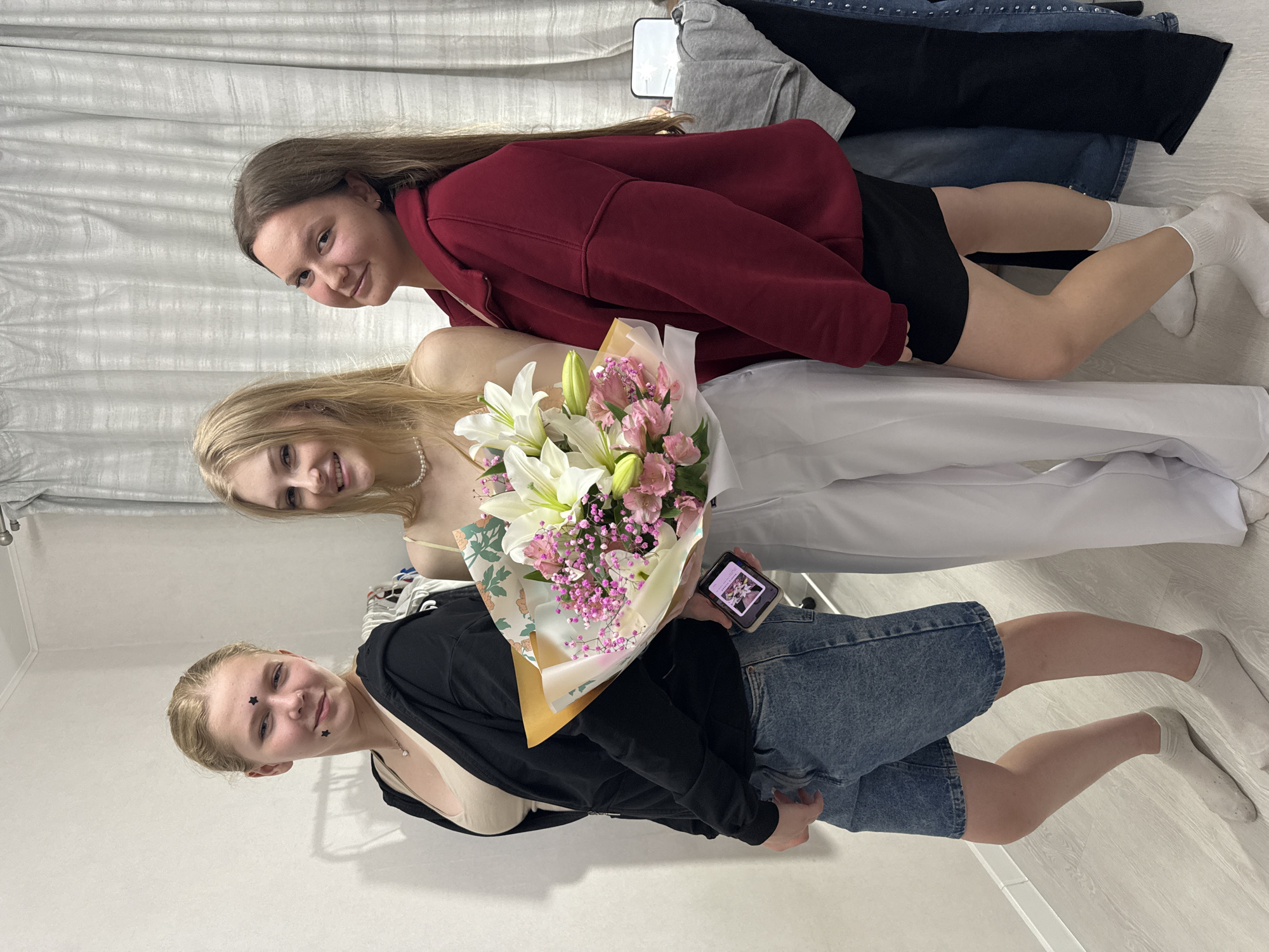 Three young women are posing for a photo. The woman in the foreground is wearing a black open jacket over a beige top and denim shorts. She has black star stickers on her face and a necklace. She is holding a bouquet of white lilies and pink flowers and looking at a smartphone. The woman in the middle is wearing a light-colored top and white pants. She has a pearl necklace and is smiling. The woman in the background is wearing a maroon hoodie and black shorts. She has long brown hair and is smiling. The background appears to be an indoor setting with curtains and a stack of clothing. The lighting suggests it is daytime.
