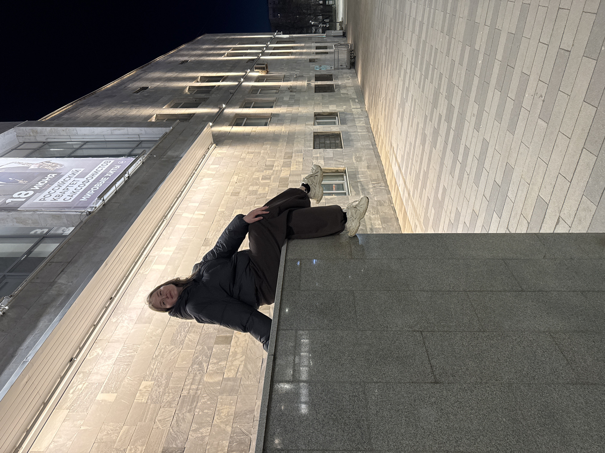 A low-angle outdoor shot at night, showing a woman sitting on a ledge against a tall building with a stone facade. The building appears to be in Perm, Russia, based on the Cyrillic text on a poster reading "18 ИЮНЯ РОССИЙСКИЙ КВАРТЕТ САКСОФОНИСТОВ МИРОВЫЕ ХИТЫ" (June 18, Russian Quartet of Saxophonists, World Hits). The woman is wearing a dark jacket and dark pants, and white sneakers. She's facing the camera with a slight smile. The building features multiple rows of windows and lights. The foreground shows a grey stone-paved area. The sky is dark.