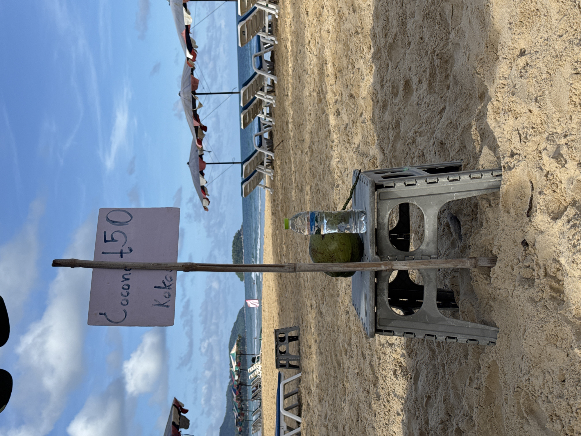 A portable grey plastic stool sits in the foreground on a sandy beach, a whole green coconut and a plastic bottle of water are placed on top of it. A sign on a bamboo pole is next to the stool, displaying "Coconut 150" and "Koko" in blue marker. The beach has lounge chairs and umbrellas lined up in the mid-ground. The ocean is visible in the background, with a small island in the distance. The sky is blue with scattered clouds. The location is Karon Beach, Karon, Thailand.