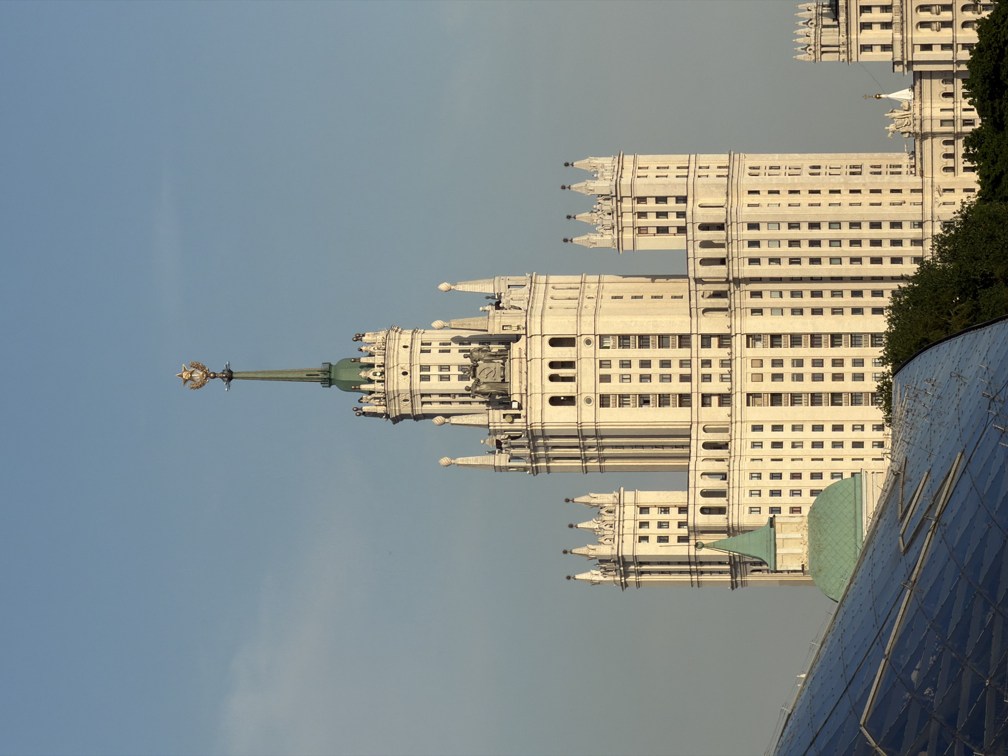 The image shows the Ministry of Foreign Affairs building, one of the "Seven Sisters" skyscrapers in Moscow, Russia. The building is white with numerous windows and ornate spires. A green spire topped with a Soviet star is visible at the top. Part of the Zaryadye Park's glass roof is in the foreground. The sky is pale blue.