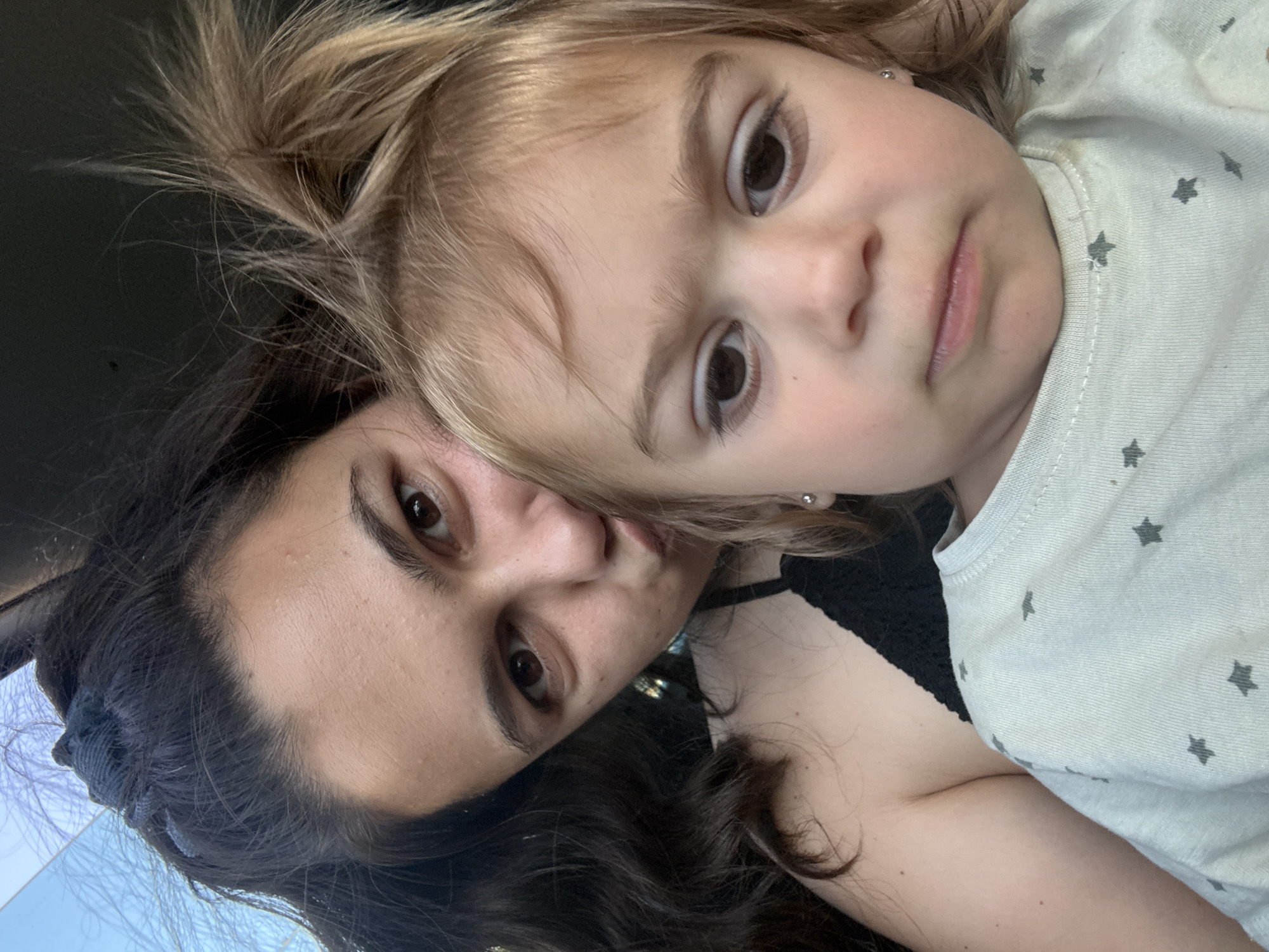 In Oreshak, Bulgaria, a close-up selfie features a woman and a young child. The woman, with dark, wavy hair pulled back, is positioned behind and to the left of the child, her face turned towards the camera with a pout. She wears a black, lacy top. The child, with blonde hair styled in pigtails, is in the foreground, facing the camera with a serious expression. The child's light-colored t-shirt, adorned with grey stars, is visible. The background is dark and out of focus.