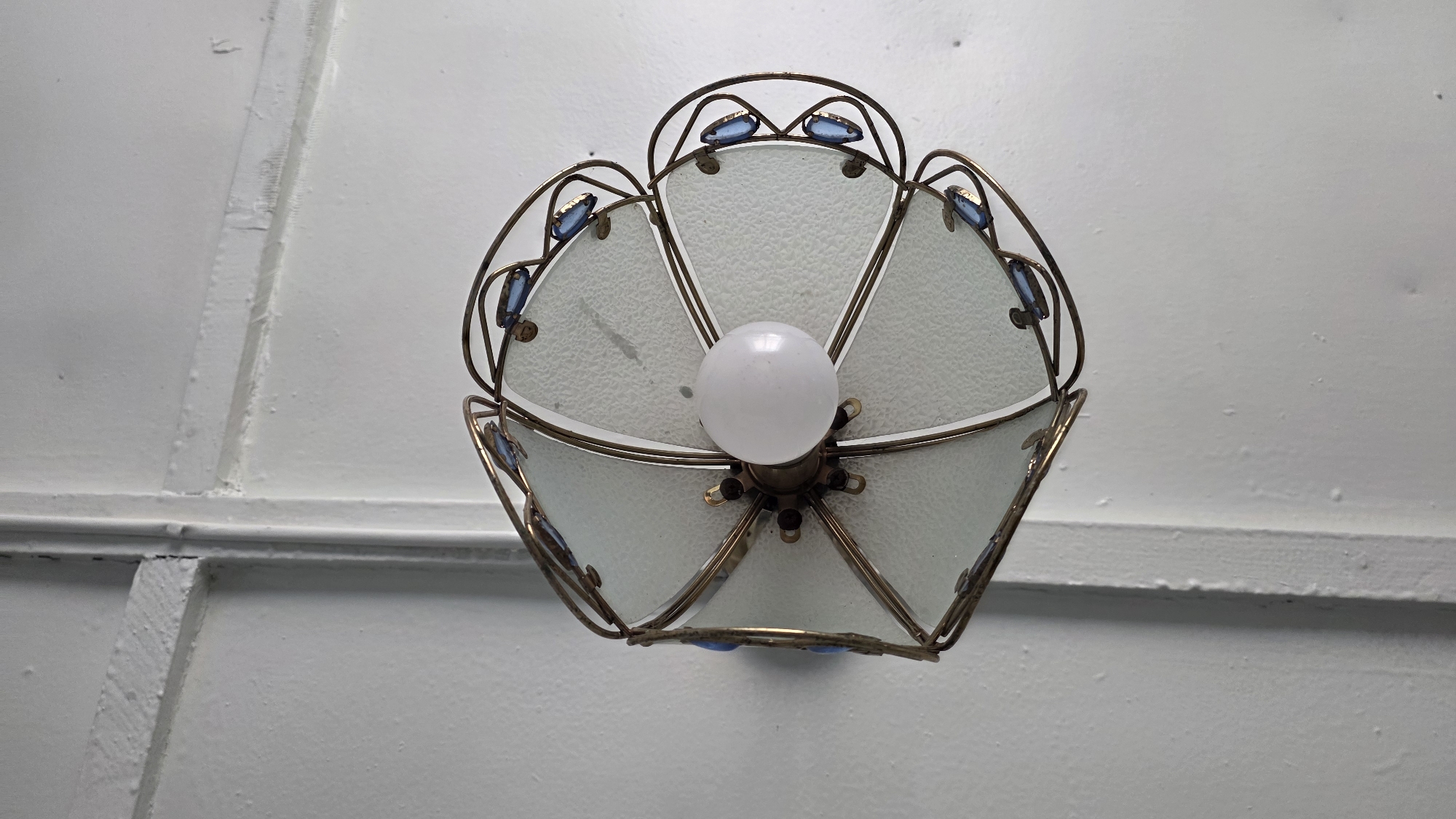 A close-up, overhead shot shows a decorative light fixture mounted on a white ceiling. The fixture has a floral design with frosted glass panels and is outlined with a thin, brass-colored metal frame. Small, rectangular blue jewels are embedded in the metalwork along the edges of the panels. A single, round, white light bulb is visible at the center of the fixture. The ceiling is painted white and features prominent, raised seams that divide it into sections. The lighting is even, and the image is sharp. The scene appears to be indoors, potentially a room or hallway. There is no information to place this image in Tokmok City, Kyrgyzstan.