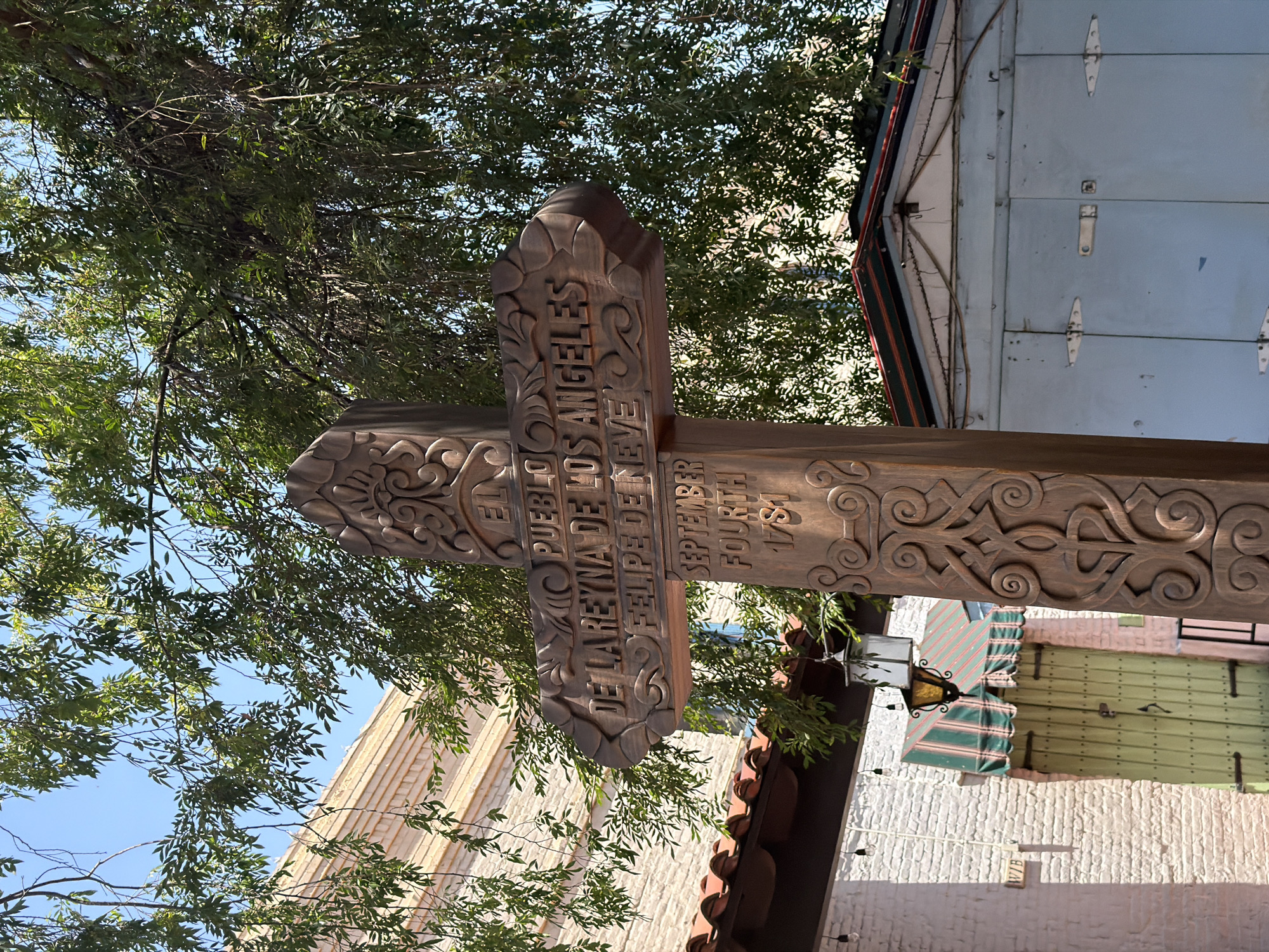 The image depicts a wooden cross-shaped sign with intricate carvings, standing against a backdrop of trees and buildings. The sign reads "El Pueblo De La Reyna De Los Angeles Felipe De Neve September Fourth 1781". The location is Los Angeles, United States of America. The buildings in the background appear to be of varying architectural styles, and the trees suggest an outdoor setting.