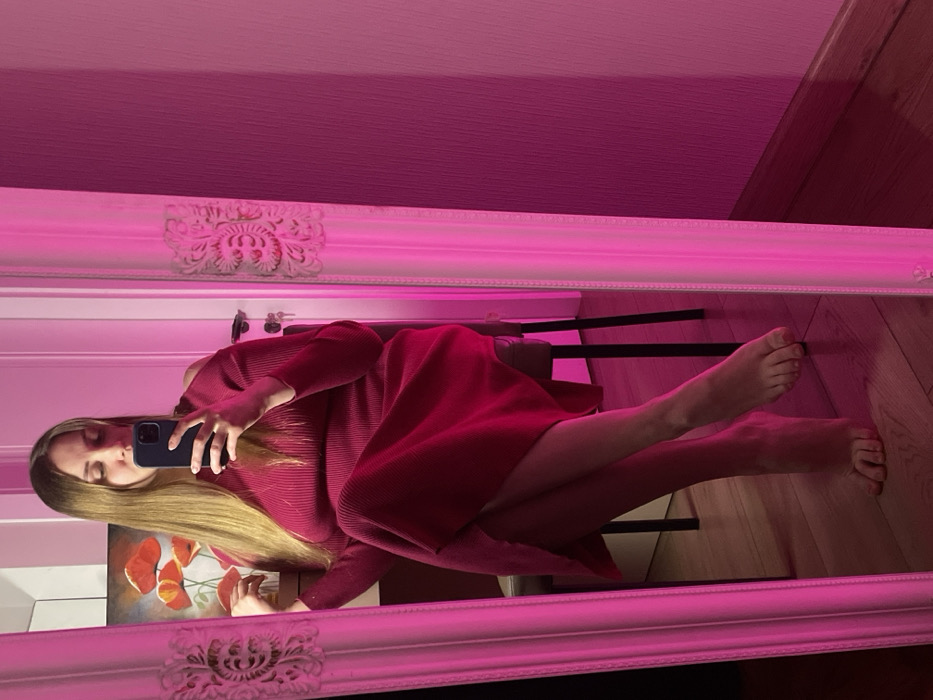 An interior scene, located in Batumi, Georgia, is depicted via a mirror selfie. The central subject is a female, visible from head to toe, with long light-brown hair, captured in the reflection of a large, white-framed mirror featuring ornate carvings. She is seated, wearing a long-sleeved, ribbed magenta dress with a cold-shoulder design and a high slit. Her bare feet are crossed at the ankles. She holds a dark-colored smartphone in her right hand, directed towards the mirror to capture the image. Her left hand rests on her left shoulder. Behind the subject's reflection, a dark chair is partially visible. The reflected background also includes a framed painting of red poppy flowers on a wall, and a white door with a key in its lock. The flooring consists of light wooden planks. The entire environment within the mirror's view is intensely illuminated by a magenta light source.