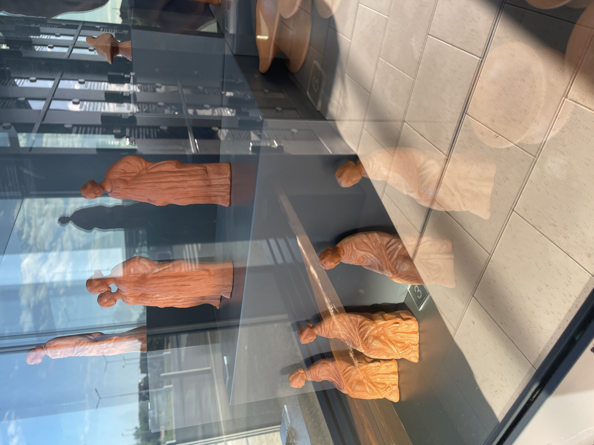 The image shows an exhibit of terracotta figures displayed behind glass, possibly in a museum or cultural center. The figures appear to be human forms, possibly representing ancient inhabitants or artifacts from the area. There are at least four rows of figures visible in the display case. The reflection in the glass shows a modern building with windows and the tiled floor. Considering the city specified is Sozopol, Bulgaria, it's likely that this exhibit is related to the ancient history and archaeology of the area, which has been inhabited since the Bronze Age and was a Greek colony known as Apollonia Pontica.
