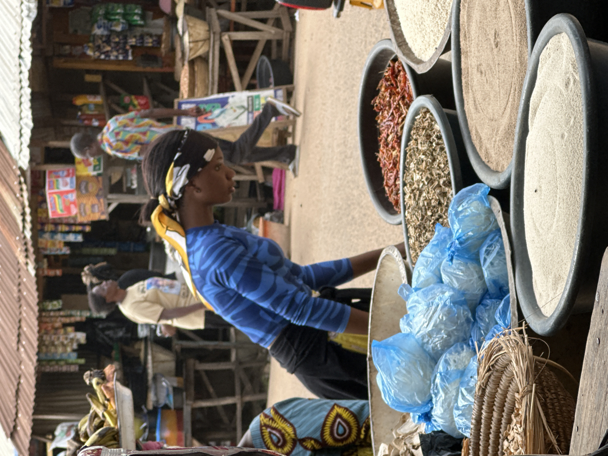 In Narayi, Nigeria, this outdoor market scene captures the bustling atmosphere of commerce. A woman in a blue, patterned long-sleeved shirt and black pants is bent over, her head down, as she appears to be organizing or displaying goods. She wears a yellow and black headscarf. In the foreground, several large, grey plastic basins are filled with various food items. One basin contains what appears to be dried red chilies, another has a mixture of chopped herbs or spices, and a third holds white grains, likely rice. A woven basket sits to the left, also filled with what looks like dried grains or seeds. Several blue plastic bags, filled with white material, are piled in a large, light-colored basin. In the background, other market stalls are visible, with shelves stocked with packaged goods and produce like bananas. People are seen in the distance, including a man in a colorful patterned shirt and a woman in a yellow shirt. The overall environment suggests a vibrant and active marketplace.